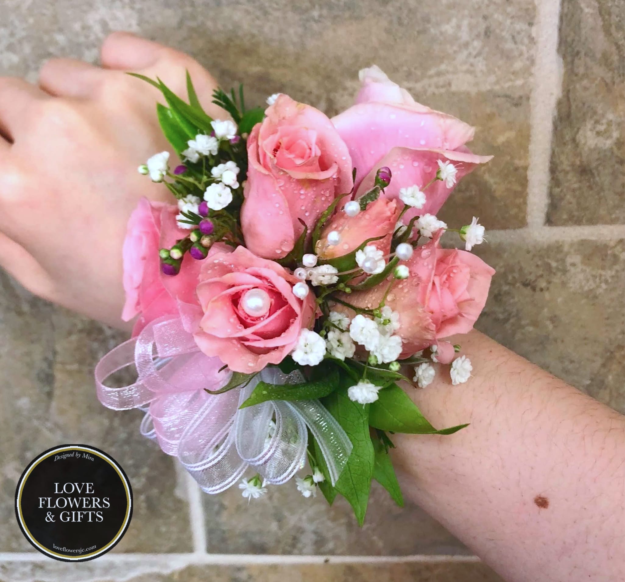 Pink rose wrist corsage with white filler flowers, pearls, and sheer ribbon on a wrist