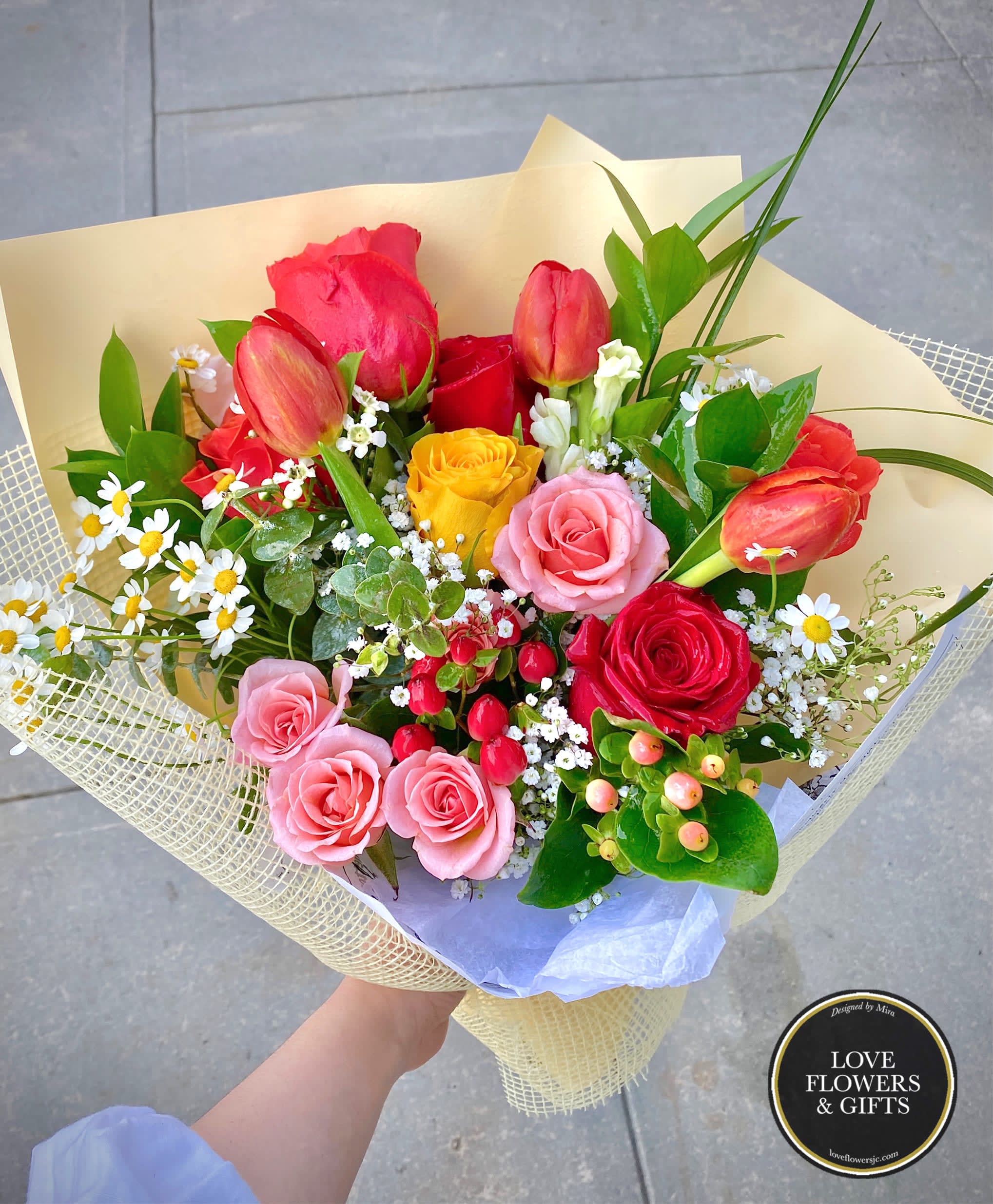 Hand-tied bouquet of mixed roses, tulips, and small white flowers wrapped in pale yellow paper