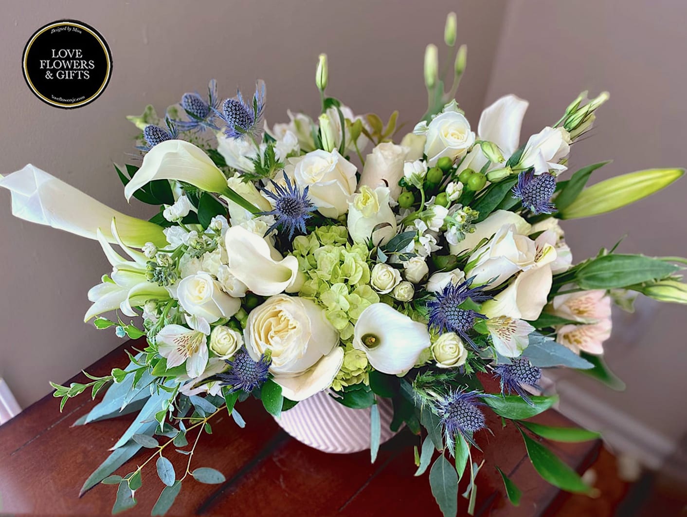 Low white and green arrangement of roses, calla lilies, and blue thistle in a white ribbed vase