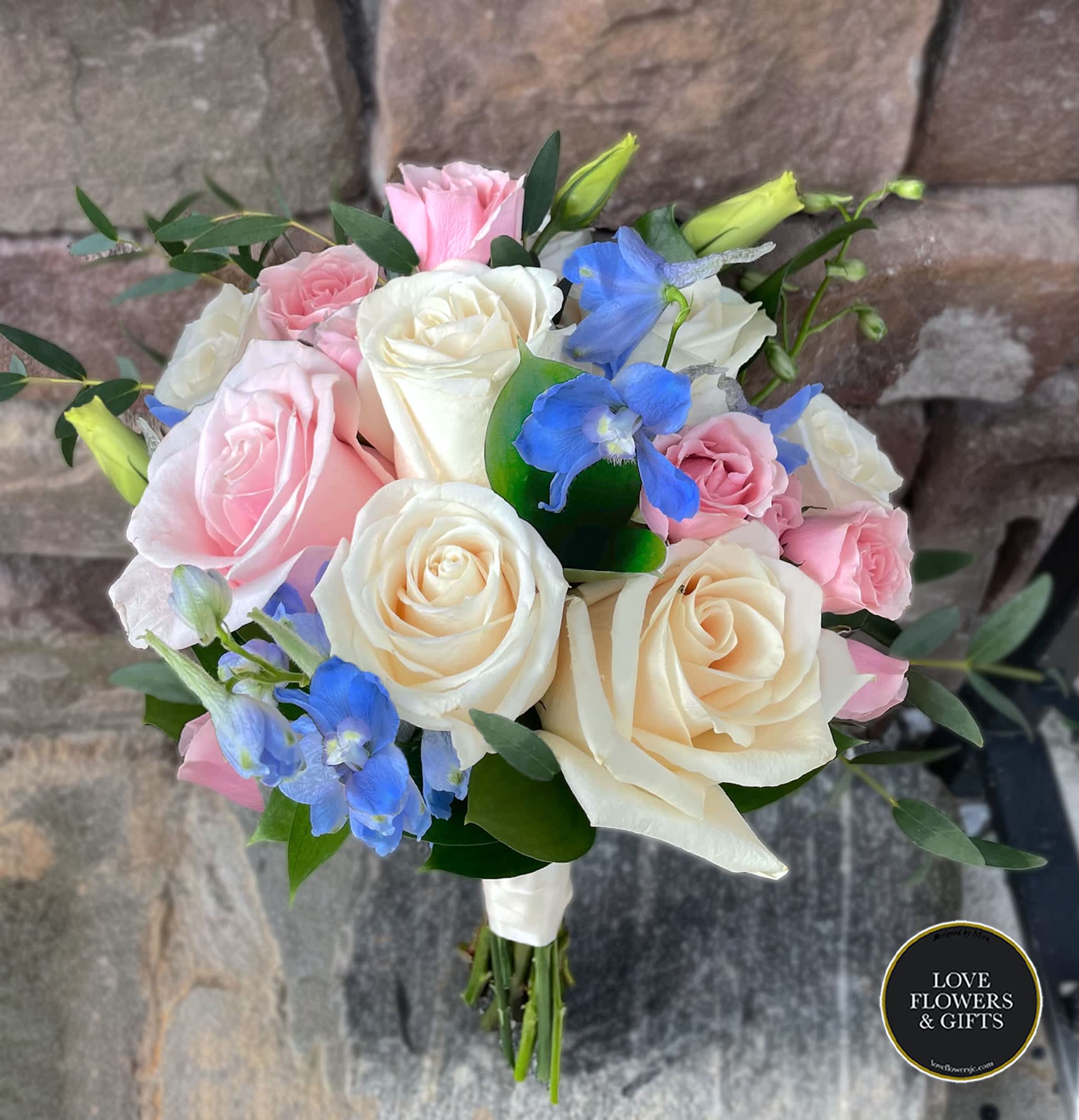 Hand-tied bouquet of ivory and pink roses with blue delphinium, wrapped in white ribbon against a stone background.