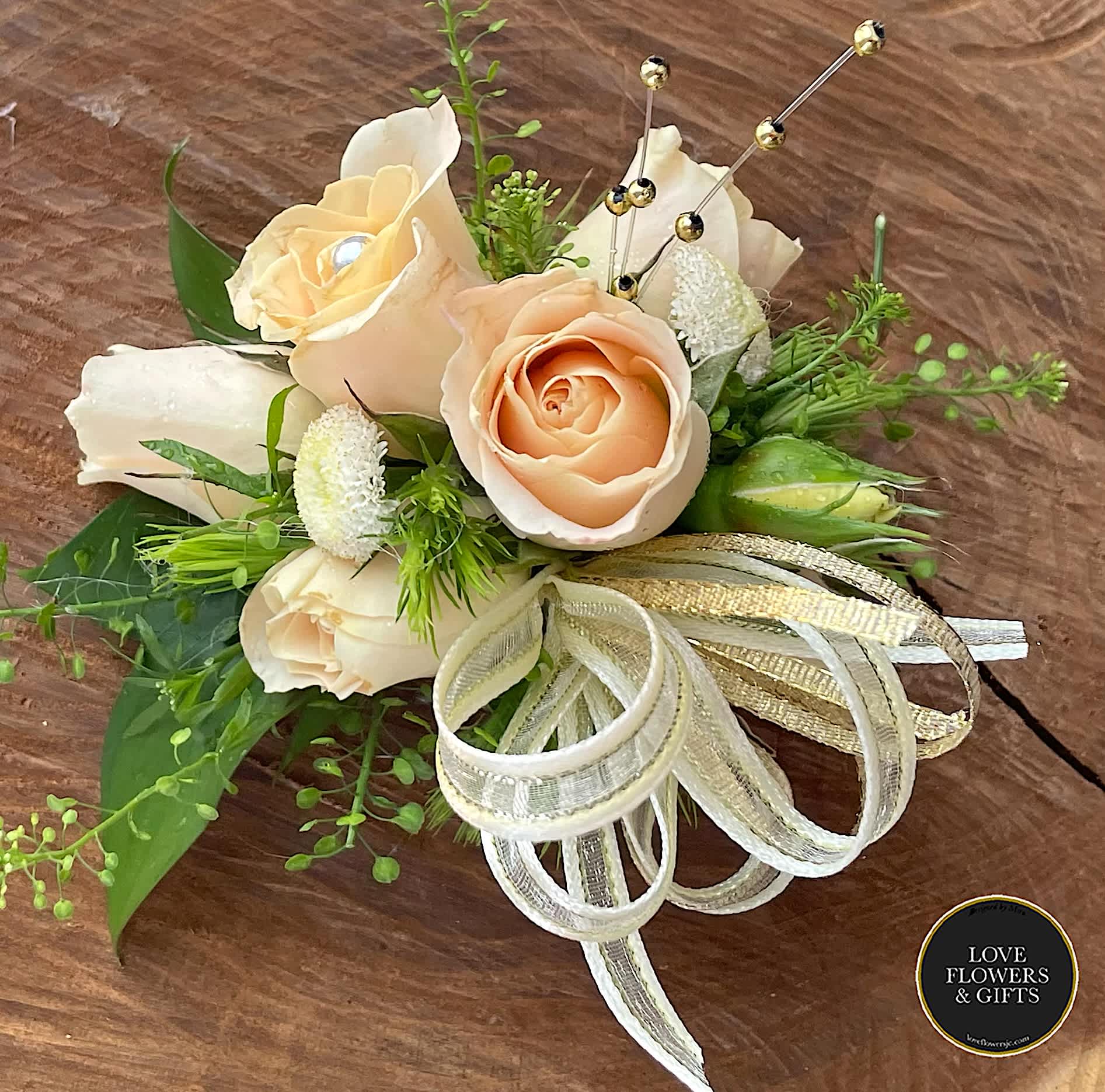 Peach rose corsage with white accents, gold ribbon bow, and jeweled pins on a wooden surface.