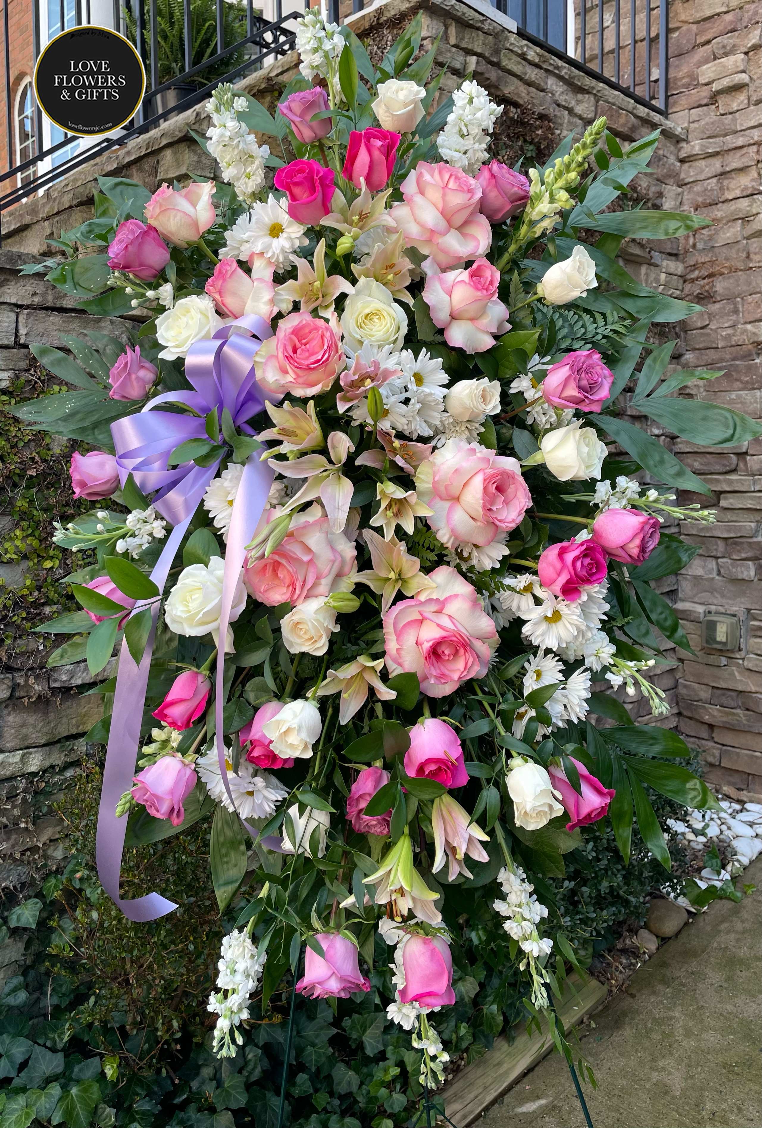 Tall standing spray of pink and white roses, lilies, and daisies with a lavender ribbon on an easel