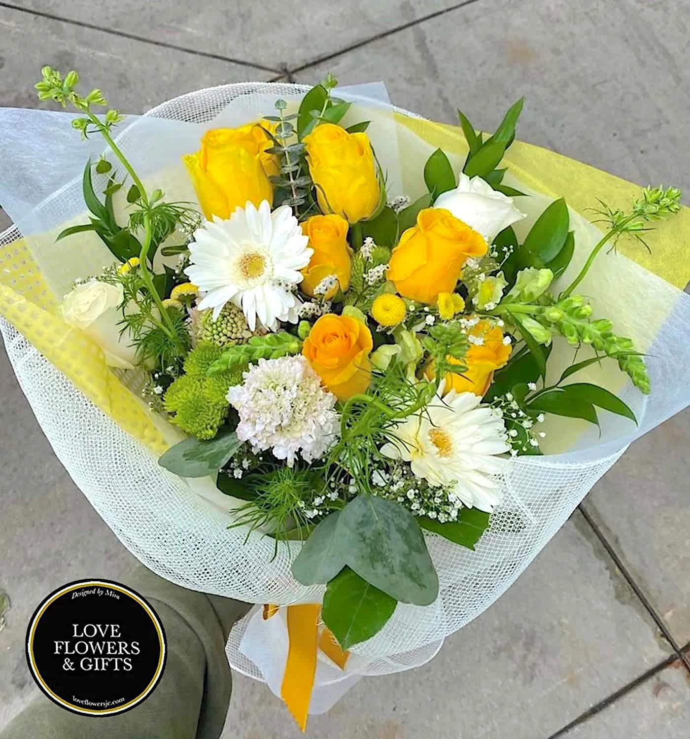 Hand-tied bouquet of yellow roses and white gerbera daisies wrapped in white and yellow mesh.