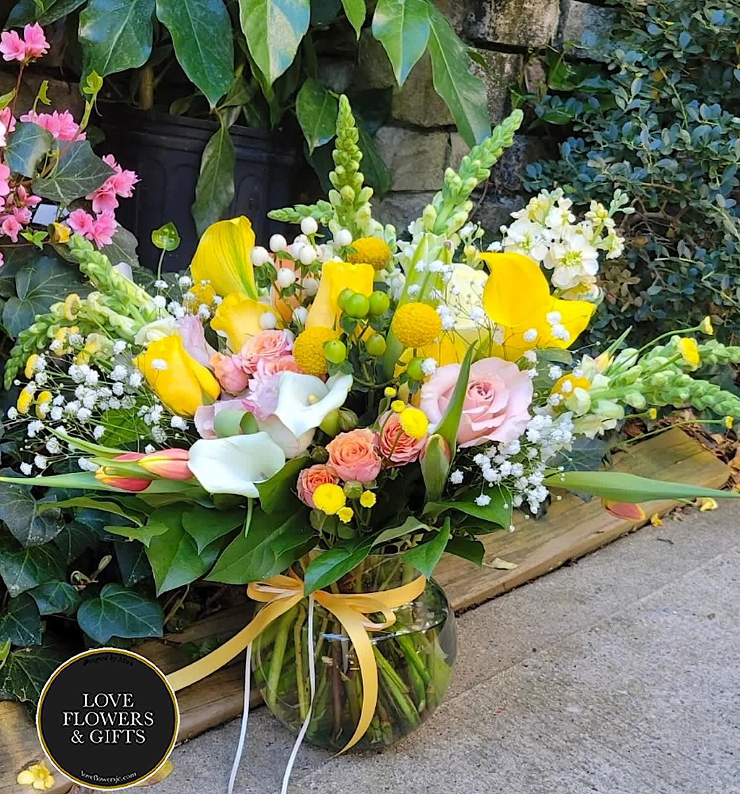 Bright bouquet of yellow calla lilies, pink roses, and white blooms in a clear glass vase with a yellow ribbon.