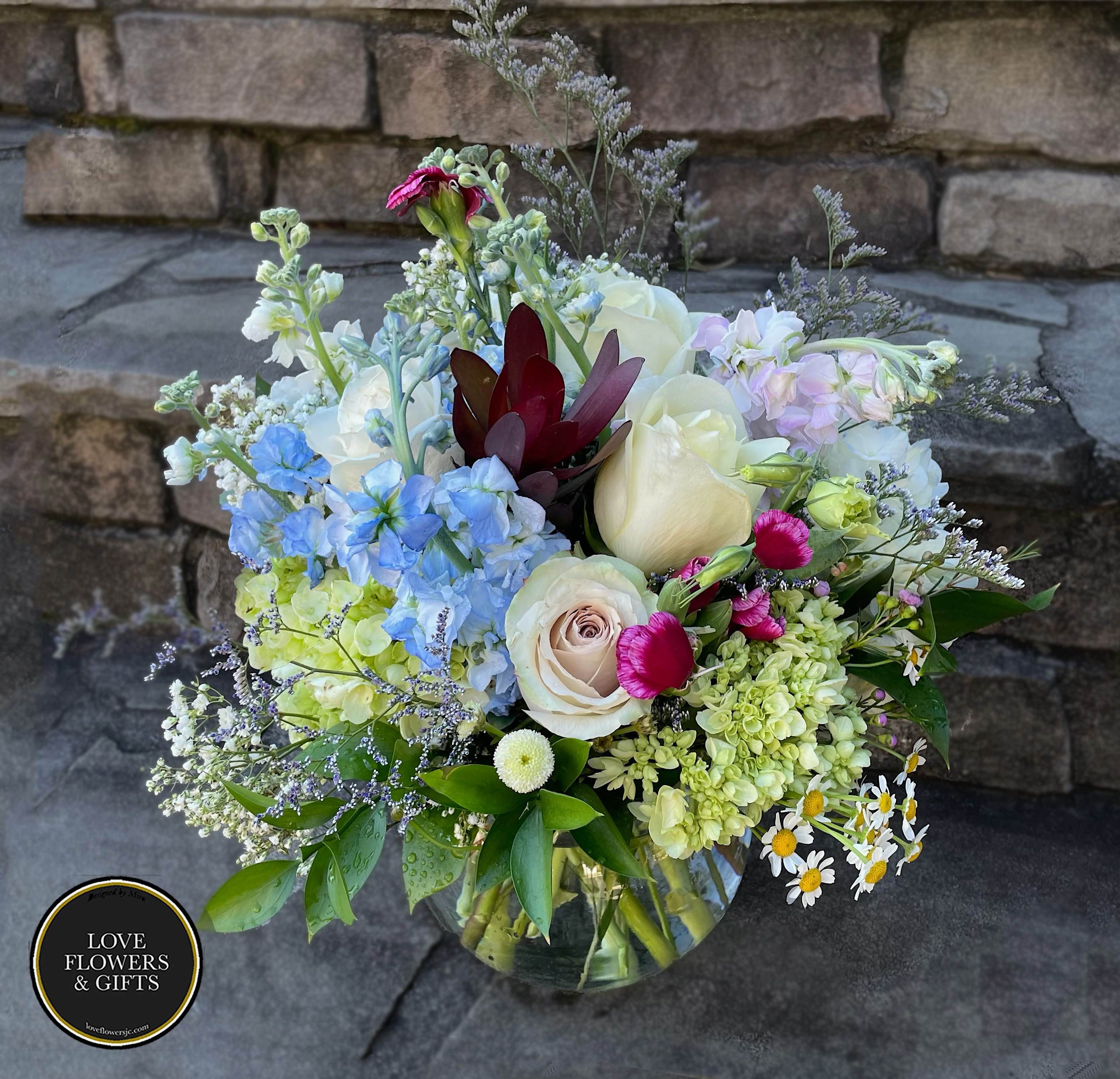 Mixed bouquet of cream roses, blue delphinium, green hydrangea and small daisies in a clear glass vase.