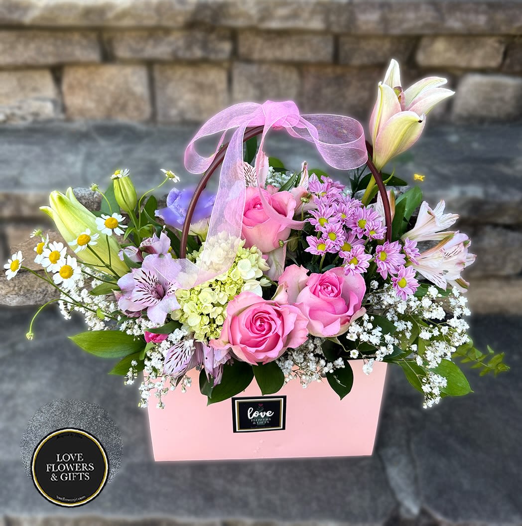 Pink gift bag of mixed flowers including pink roses, lilies, daisies, and filler with a sheer pink bow.
