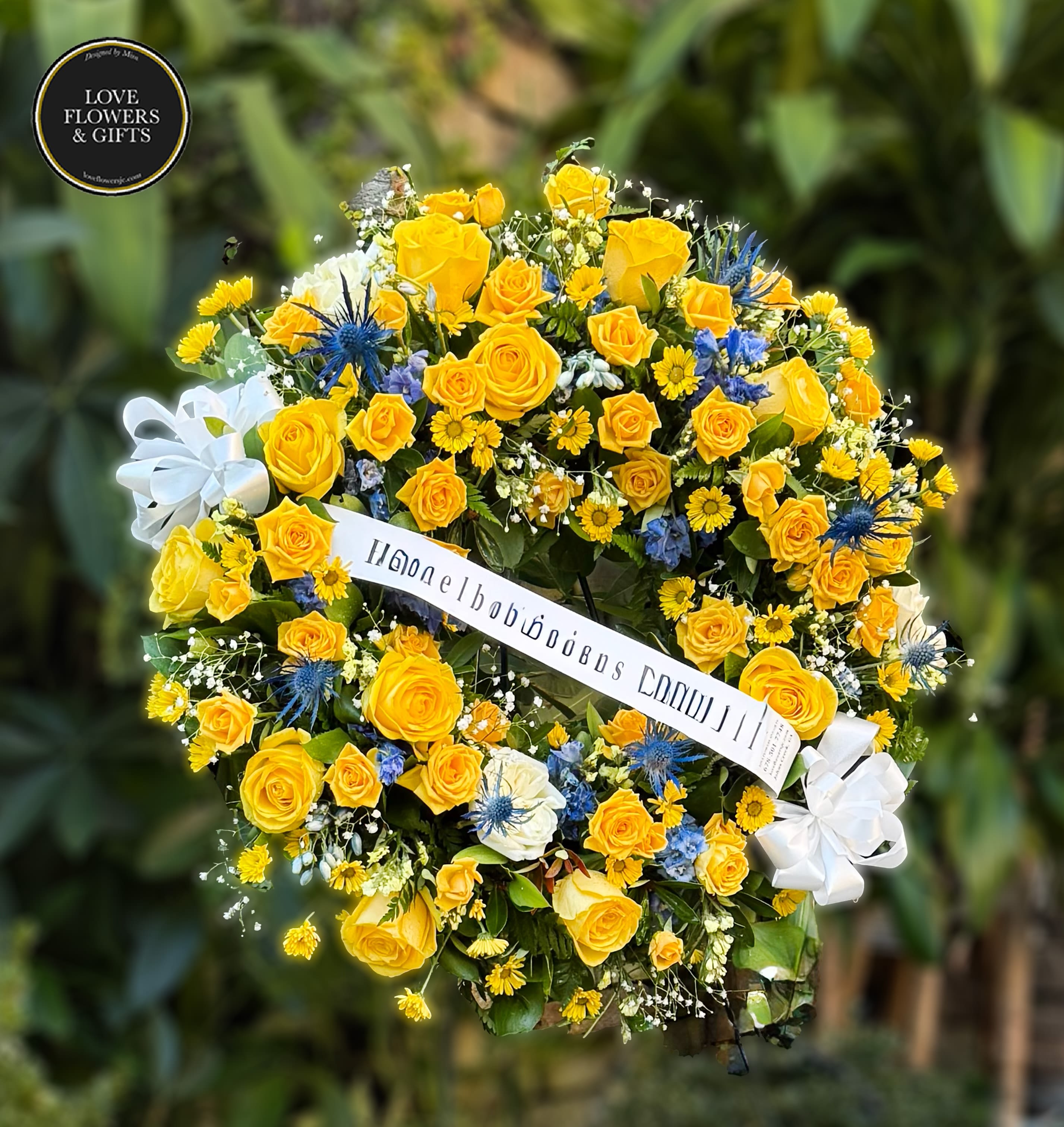 Round standing wreath of yellow roses and daisies with blue accents and a white ribbon banner.