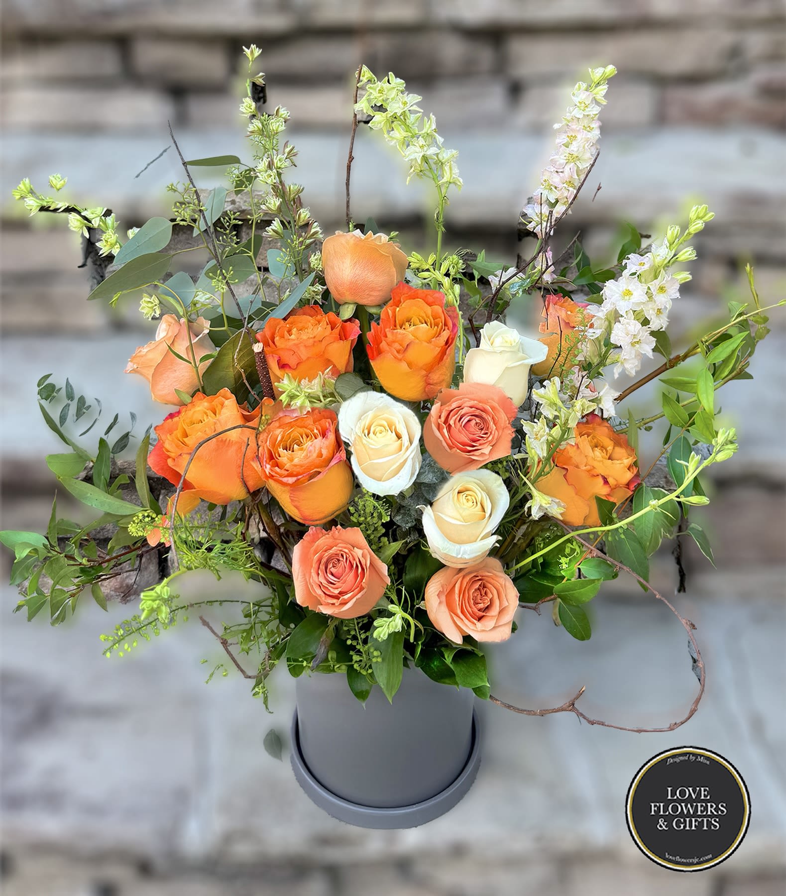Arrangement of orange and cream roses with tall white blooms in a gray cylinder vase