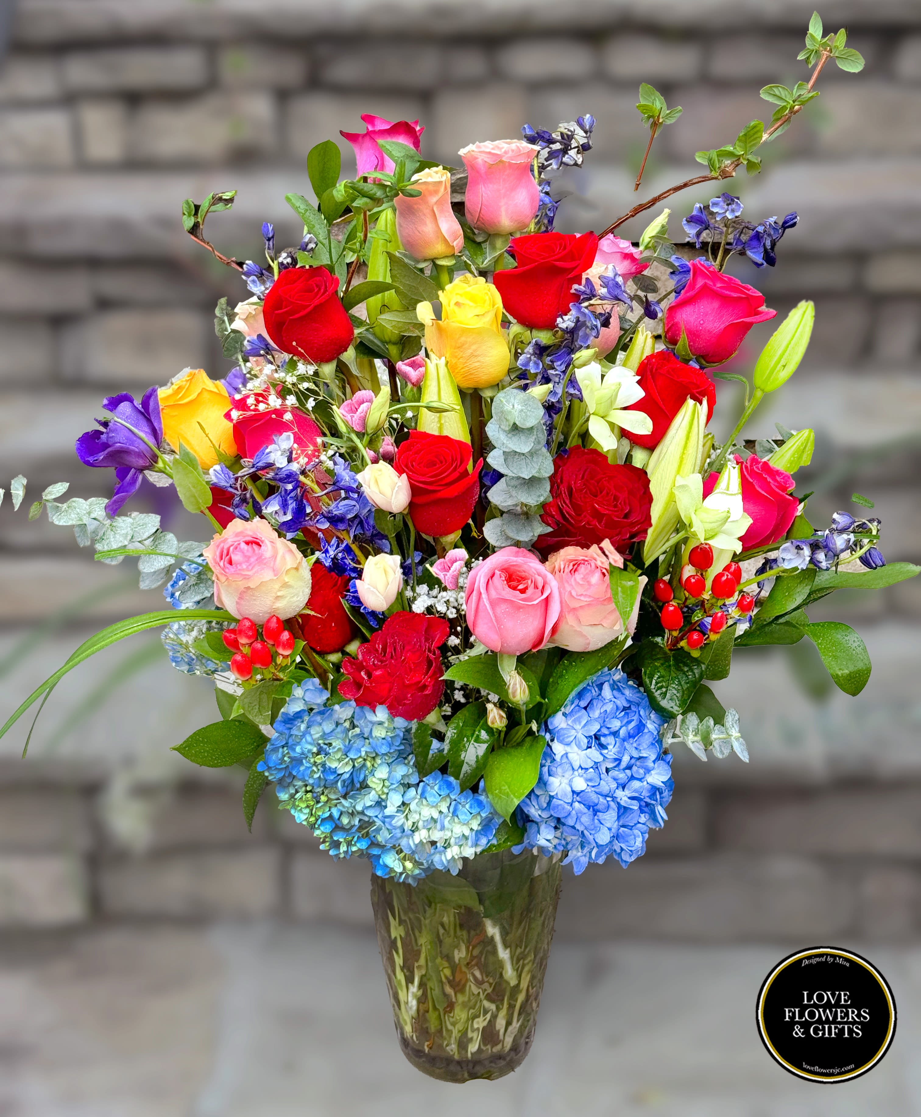 Tall mixed bouquet of multicolored roses, lilies, irises, and blue hydrangeas in a glass vase
