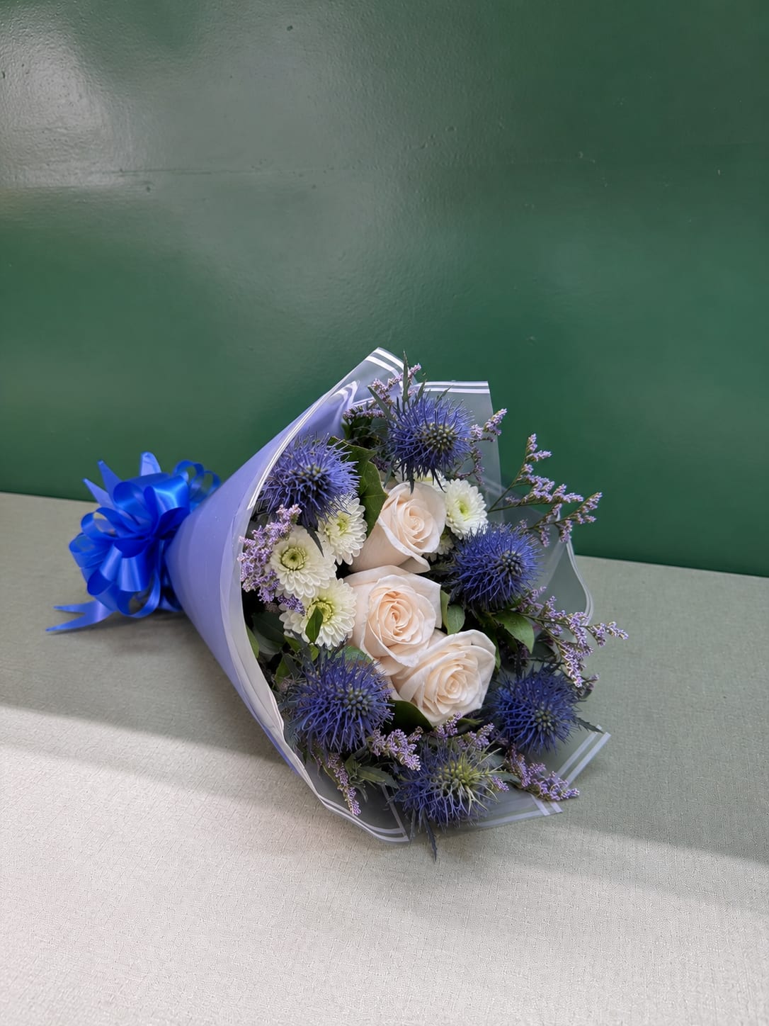 Bouquet of pale roses, white blooms, and blue thistle wrapped in lavender paper