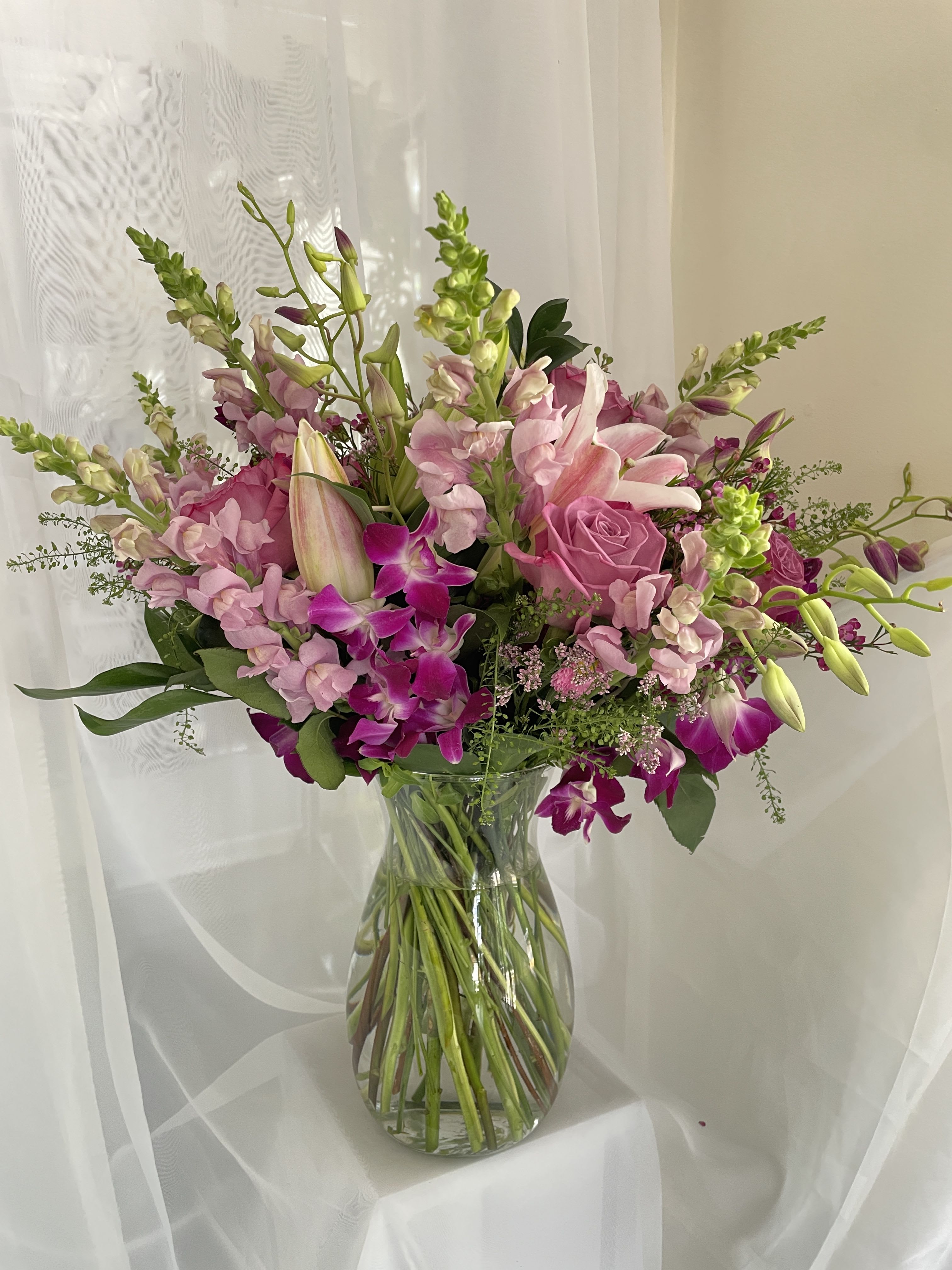 Paradise of Purples Arrangement - Bright magenta orchids and lavender roses mingle with tall snapdragon spikes to create a romantic, garden-style bouquet-perfect as a housewarming gift or a love-and-romance statement. This flower arrangement for Get Well wishes or Valentine's Day offers an airy yet lush silhouette that will make the person you're thinking of feel seen and celebrated, whether you're marking an anniversary, Women's Day, or simply brightening a bedside or living room.  Layered at varied heights in a clear glass vase, the Paradise of Purples arrangement mixes dendrobium orchids, lilies, snapdragons, roses and complementary greens for texture and movement. Colors range from deep purple to pink, white and fresh green, with upright stems adding vertical drama and softer blooms creating a full, feminine feel. The vase gives it a clean, modern finish that fits well on a dining table, entry console, or hospital bedside. 