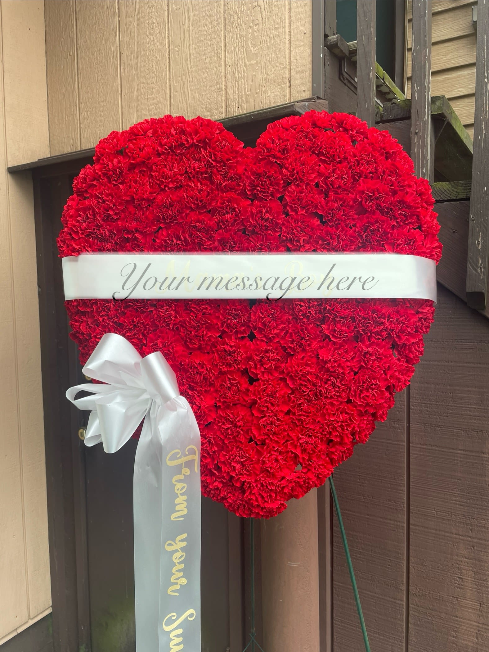 Solid Red Heart - A lush heart of densely packed red carnations gives this flower arrangement the meaning of love. A wide white satin message band crosses the center so you can add a personalized note, and a white bow with a long trailing ribbon hangs from the lower left for a graceful finishing touch. The arrangement is crafted entirely from red carnations with white ribbon accents, creating a clean two-color palette that reads elegant and heartfelt up close or from across the room.  Choose this solid red heart to send your condolences.