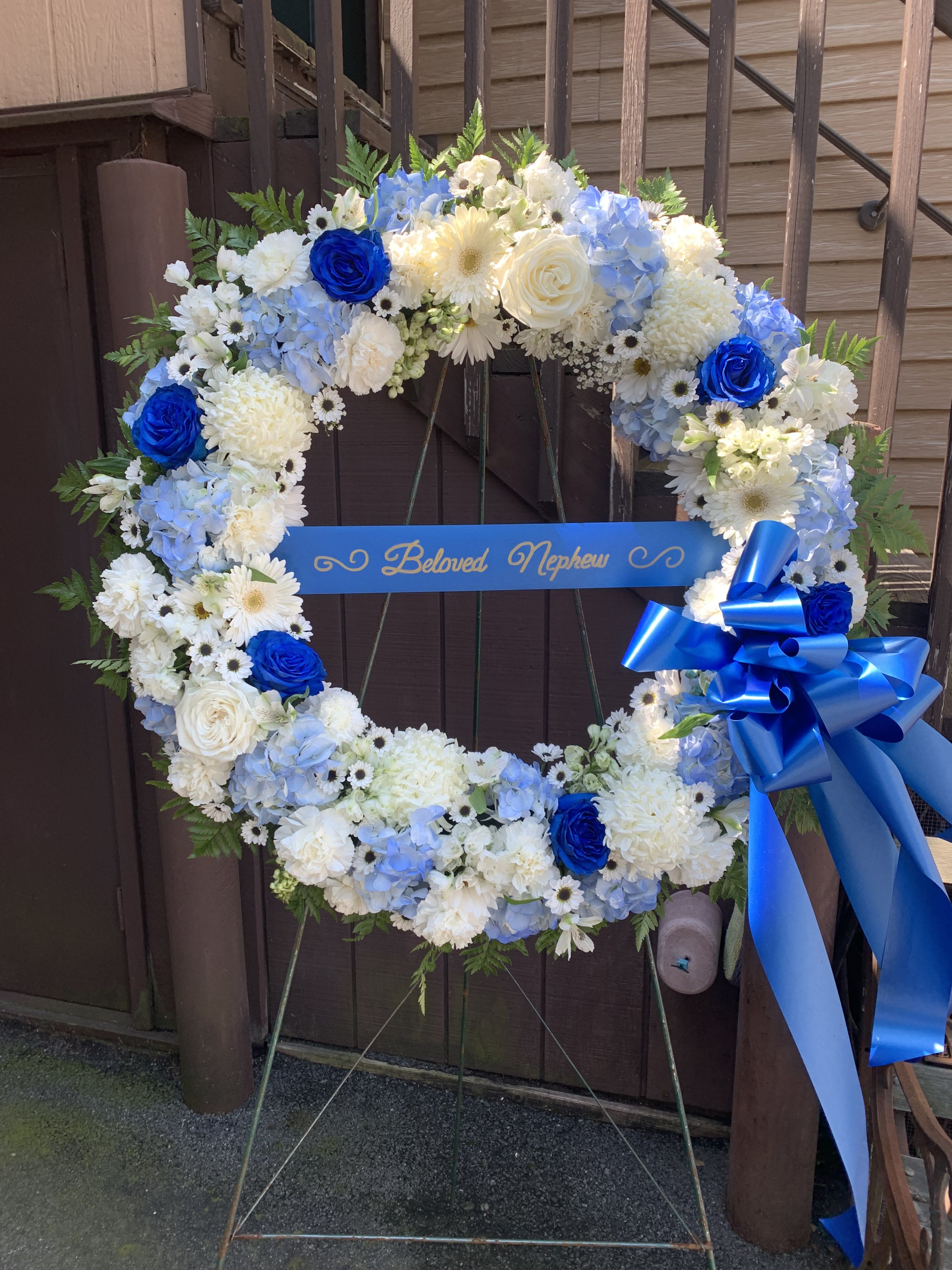 Sympathy Wreath White and Blue - A large circular sympathy wreath in serene white and blue offers a formal, comforting tribute for funeral or remembrance services. The cool palette-crisp whites, soft powder blues, and striking royal blue roses-creates a peaceful, respectful impression that helps the person you're honoring feel remembered and dignified.  Densely packed carnations, chrysanthemums, hydrangeas, roses, and small daisy-like filler flowers form the ring, with fern greenery woven through for texture and contrast. A bright blue ribbon bow and a horizontal name plaque cross the center for a traditional memorial presentation; the wreath is arranged to sit upright on a wreath easel for display.  