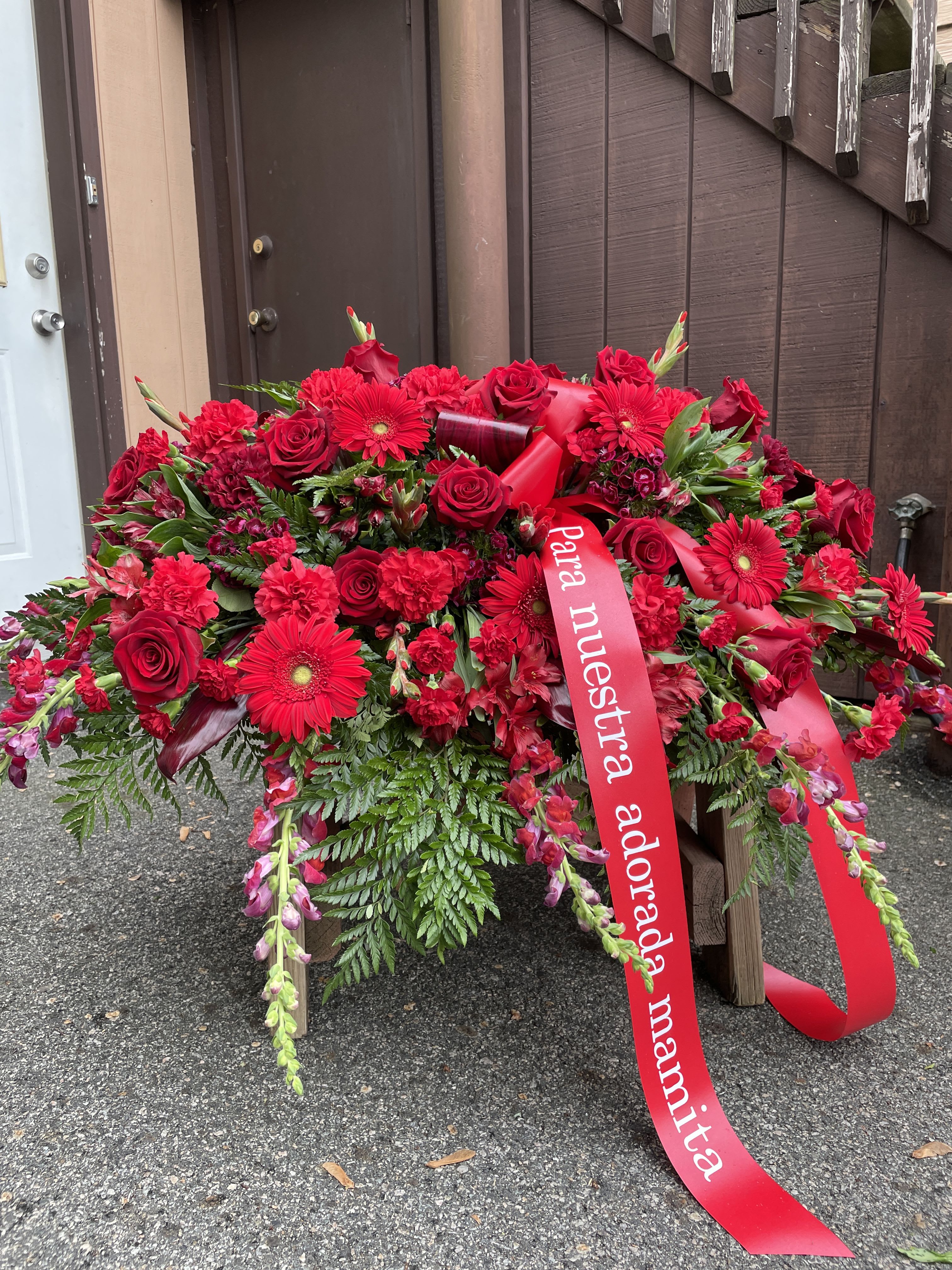 Casket Spray Endless Love - This large, broad spray features layered clusters of roses, carnations, and trailing red alstroemeria accented by upright snapdragon spikes and fern-like greenery for a full, cascading silhouette. The composition sits on the casket and includes a long red ribbon with printed text at the center, giving the piece a formal, commemorative focal point. The dense, low profile and generous dimensions ensure it reads clearly from the front of a service, offering a strong visual statement.  Perfect for a funeral service or graveside tribute, this arrangement conveys enduring love and respect-an appropriate choice for a close family member or dear friend. The flowers are arranged by our florists for lasting presentation; if you need specific ribbon wording, delivery timing, or positioning on the casket, please let us know when ordering so we can accommodate your wishes.