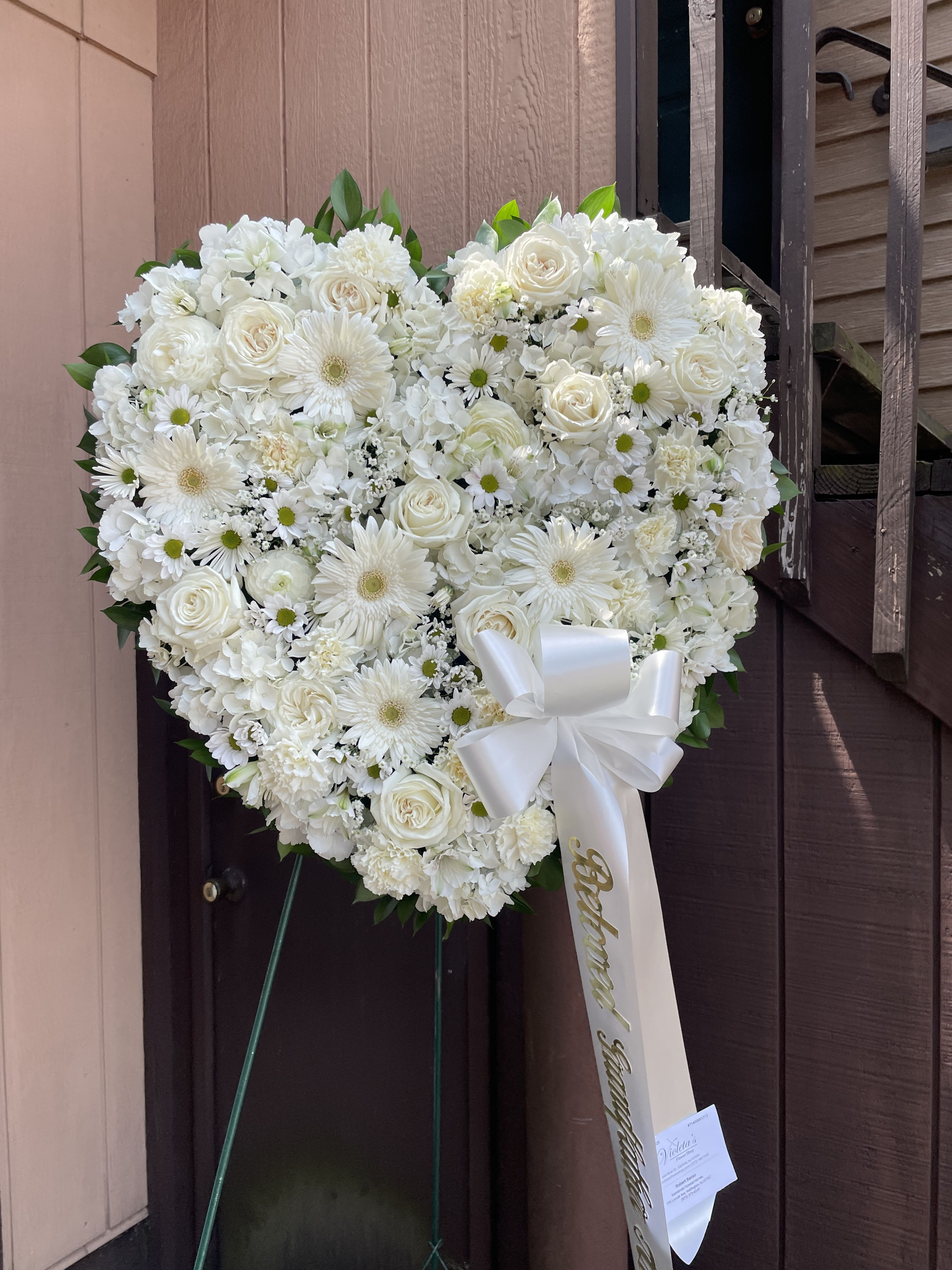 Solid All White Heart - A large heart-shaped standing spray in an all-white palette creates a calm, respectful statement perfect for a sympathy or funeral tribute. The solid white heart arrangement uses roses, gerbera daisies, small daisy blooms, carnations, and clusters of hydrangea to form a dense, symmetrical shape that reads as both elegant and consoling - the kind of arrangement that helps the person you're honoring feel cherished and remembered.  This arrangement is mounted on a standing easel and finished with a wide white ribbon bow and a card for your message. A soft green edge peeks through around the perimeter, adding subtle contrast and a gentle, natural outline to the white blooms. The composition is compact and formal, designed to hold its shape during service and viewing.