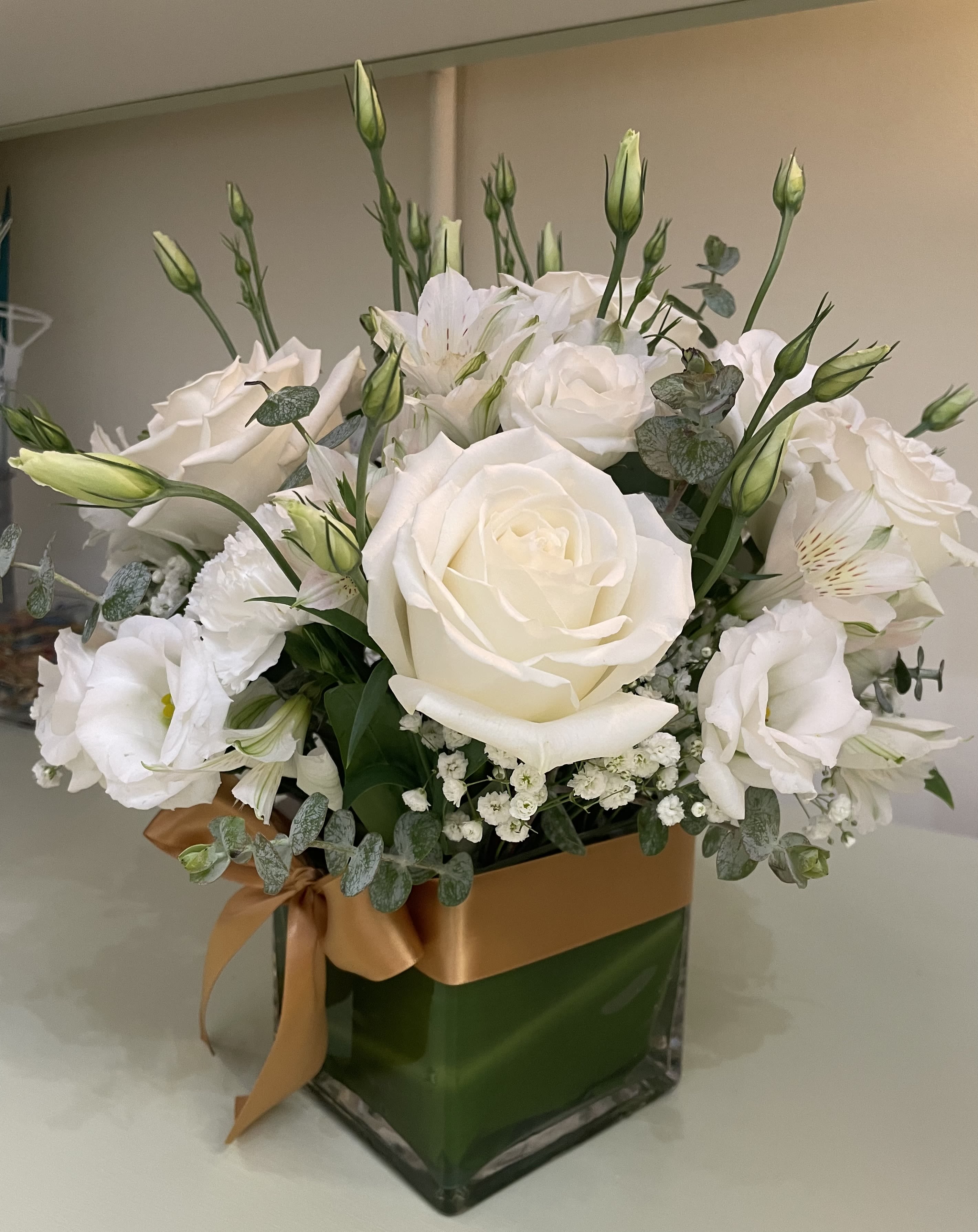 Roses and Lisianthus Arrangement - White roses take center stage in this elegant sympathy arrangement, perfectly suited for a funeral tribute, wedding table, or any formal occasion where a calm, refined presence is needed. The upright, airy design and pale green foliage give the bouquet a serene feeling-an easy way to show someone they're being held in gentle thought.  Surrounding the roses are white lisianthus and delicate white filler blooms that create soft layers and texture, with a few unopened buds adding natural height and freshness. The flowers sit in a clear square glass vase tied with a tan satin ribbon, giving a clean, modern silhouette that reads as both understated and intentional. Ideal for sending condolences, marking a milestone, or bringing quiet beauty to a reception, this arrangement arrives ready to display; place in a cool spot away from direct sunlight and refresh the water every two days to keep blooms looking their best.
