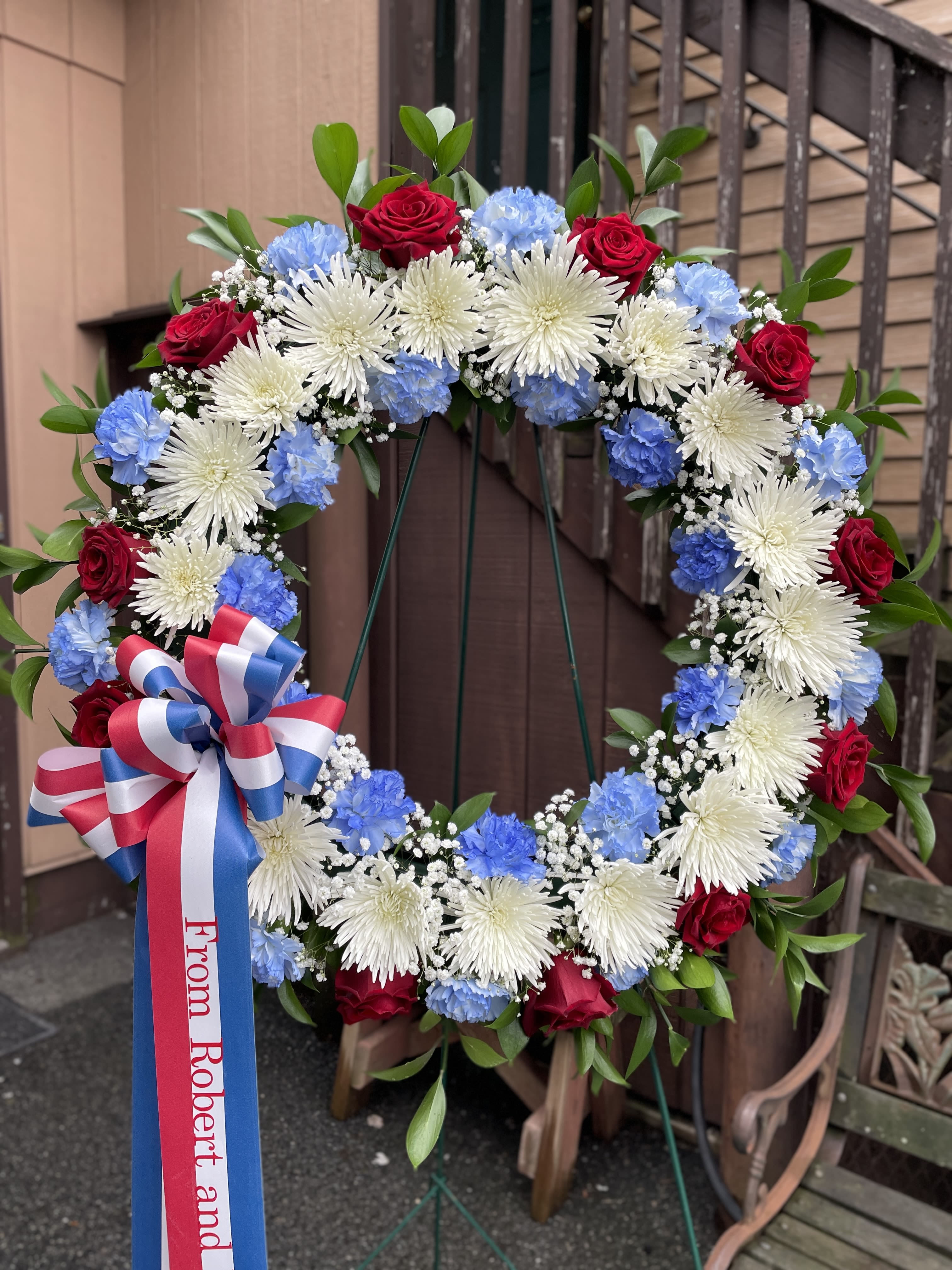 Patriotic Wreath - A large circular wreath arranged with red roses, white spider chrysanthemums, and pale blue carnations-this patriotic wreath makes a respectful statement for memorials, Veterans Day, or any commemorative occasion. The dense ring of blooms and the red, white, and blue ribbon bow create an immediate visual of honor and remembrance that the person you're thinking of will recognize and appreciate.  Ideal for graveside tribute, memorial services, or flag-raising ceremonies, this arrangement conveys solemn respect while still feeling hopeful and dignified. Care: made to be displayed upright on the included stand; keep out of direct heat and mist lightly if needed to maintain freshness during event use.