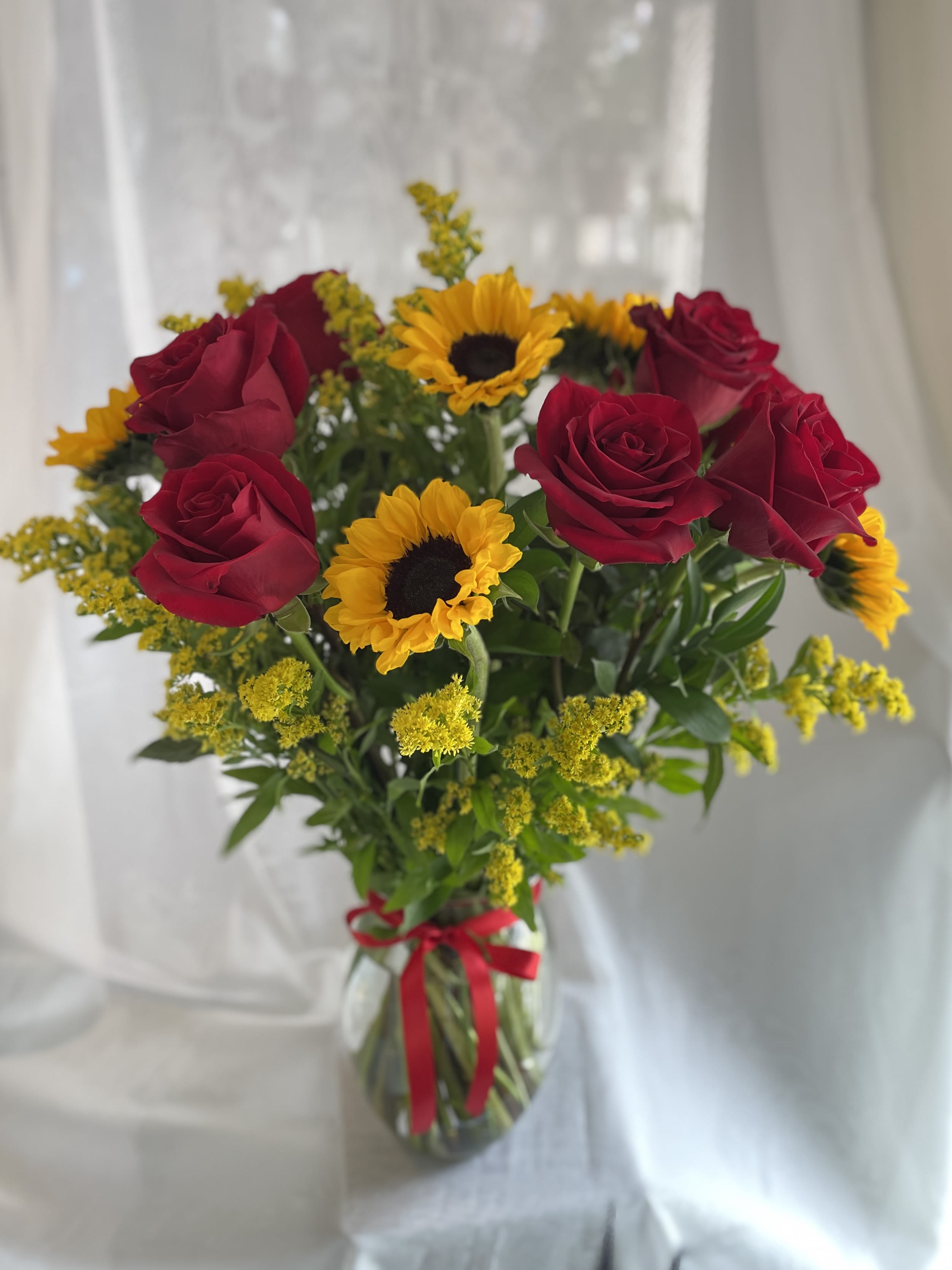 Roses and Sunflowers Arrangement - Striking deep red roses paired with sunny yellow sunflowers create a bold red-and-gold contrast that feels both romantic and celebratory - perfect as a birthday bouquet, anniversary flowers, or a cheerful "just because" surprise for the person you're thinking of. The warm mix of colors reads joyful and heartfelt, the kind of arrangement that makes someone feel truly noticed the moment it arrives.  This flower arrangement features rich red roses, bright yellow sunflowers, and small yellow filler blooms set against fresh green foliage. The stems are gathered in a clear glass vase tied with a red ribbon for a simple, gift-ready presentation that looks elegant on a dining table or desk. Ideal for everyday gifting, it brings vibrant color and a sunny scent to any room.