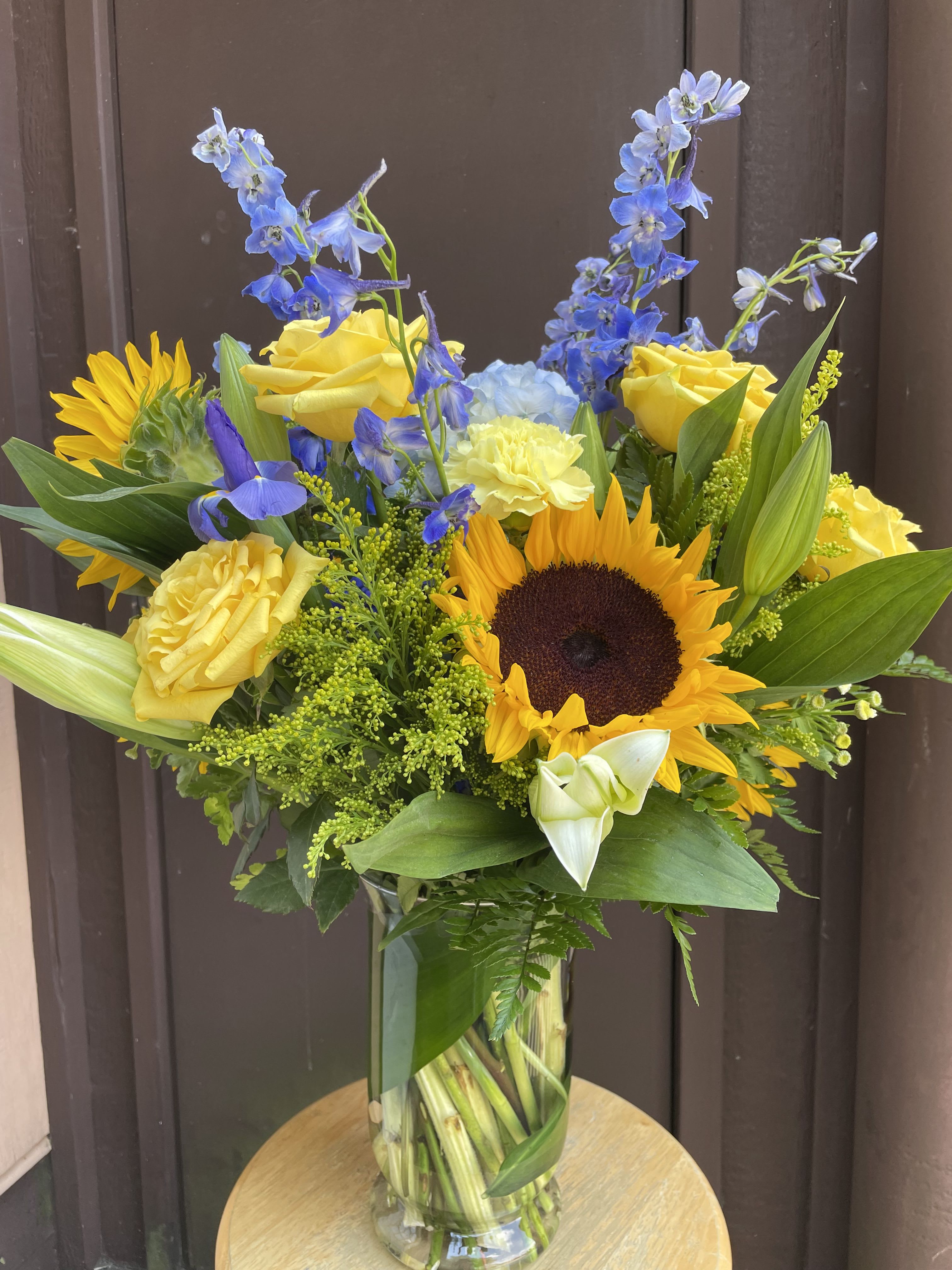 Summery Blooms Arrangement - Bright yellow roses and sunflowers meet tall blue delphinium spikes in this Summer Sky flower arrangement, creating a striking birthday bouquet or summer celebration centerpiece that reads joyful and energetic at first glance. The cool blue and warm yellow contrast makes a bold visual statement while white mini calla lilies and pale carnations soften the composition-perfect for a thank-you gift or for brightening a special anniversary or hostess moment.  Expect a mix of roses, sunflowers, delphinium, iris, carnations, and mini calla lilies in yellow, blue, green, and white tones; the tall delphinium spikes add height and the sunflowers bring sunny focal points, while the mini callas and pale blooms add elegant texture.  