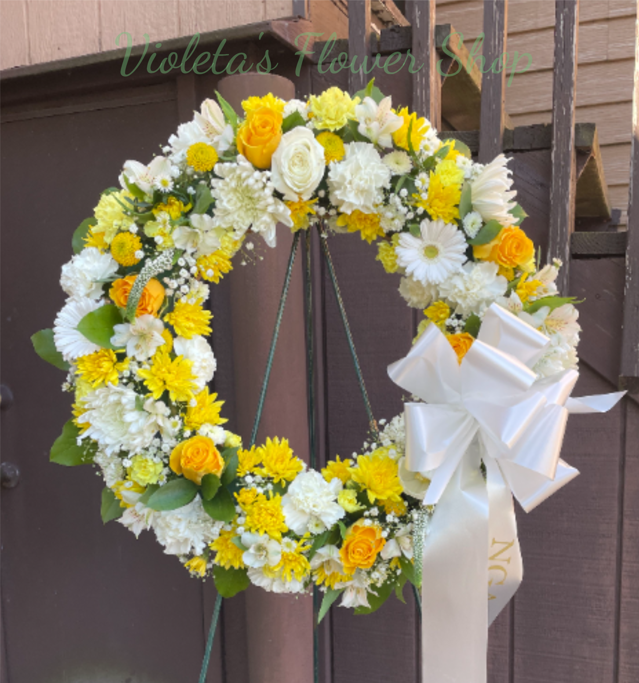 Golden Life Wreath - A bright circular wreath that reads as both celebratory and respectfully composed-perfect as a sympathy tribute or a sunny arrangement for congratulations. The Golden Life wreath pairs white and yellow blooms to create a fresh, uplifting look: white roses and chrysanthemums provide a calm, elegant base while yellow daisies and alstroemeria add cheerful highlights that catch the eye.   Densely packed roses, chrysanthemums, daisies, carnations and select filler blooms are arranged on a traditional floral wreath form and finished with broad green leaves for structure. A large white satin ribbon bow is attached to the right side and the wreath comes ready to display on the included floral wreath stand. Its full, balanced composition and polished satin ribbon give the recipient the sense of care and dignity you want to convey, whether you're celebrating a life, offering comfort, or marking a joyful occasion.