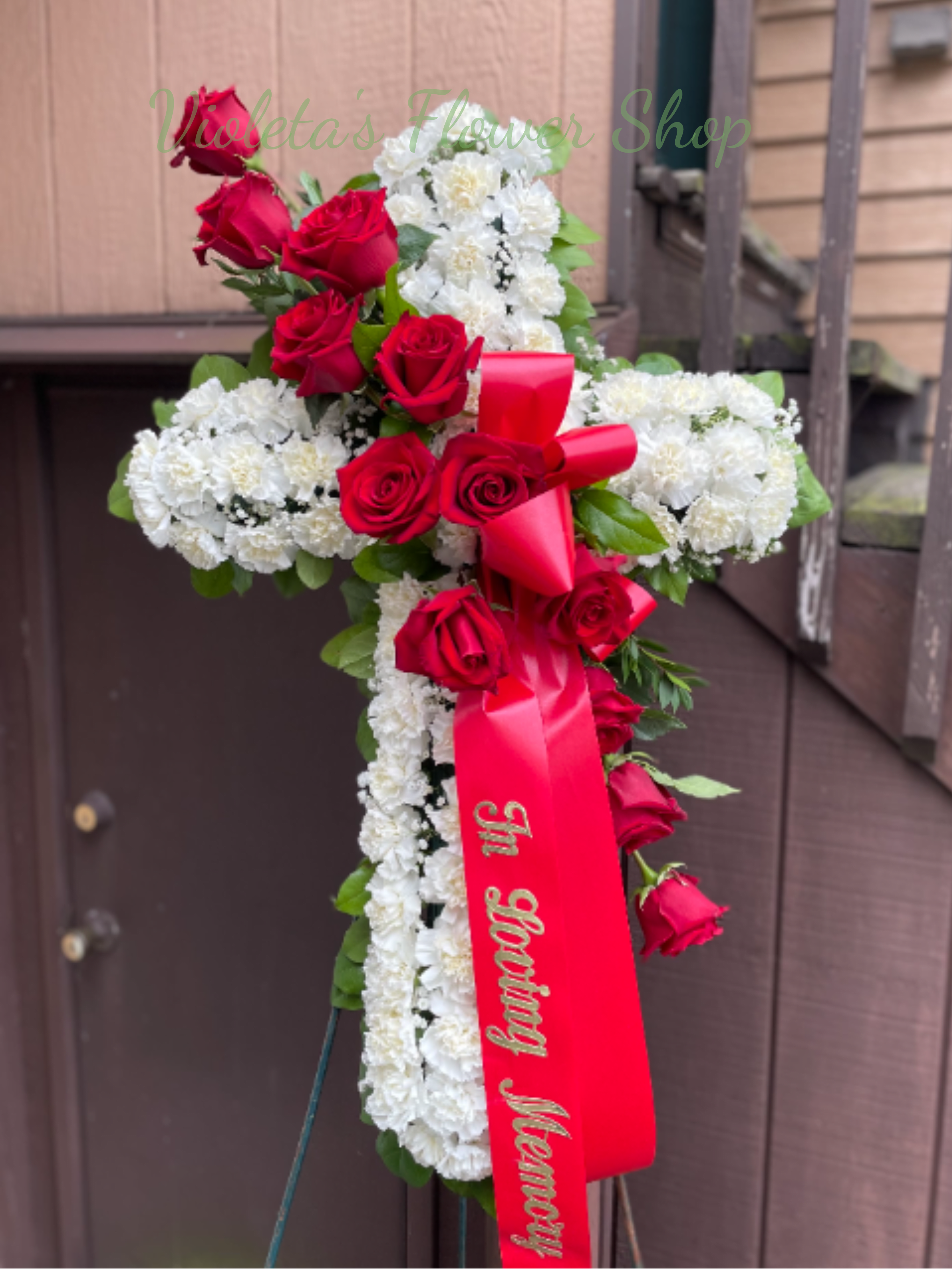Bless Forever Cross - A dignified standing sympathy cross of white blooms with bold red accents, designed as a funeral or memorial tribute that reads as both reverent and unmistakably devotional. The cross shape is densely covered in white flowers (roses or carnations depending on the piece) so the surface reads luminous and textured; clusters of red roses are placed across the upper and front sections to draw the eye and convey love and respect. Black or red satin ribbon-sometimes bearing gold "In Loving Memory" lettering-drops down the center for a formal, traditional finish.  