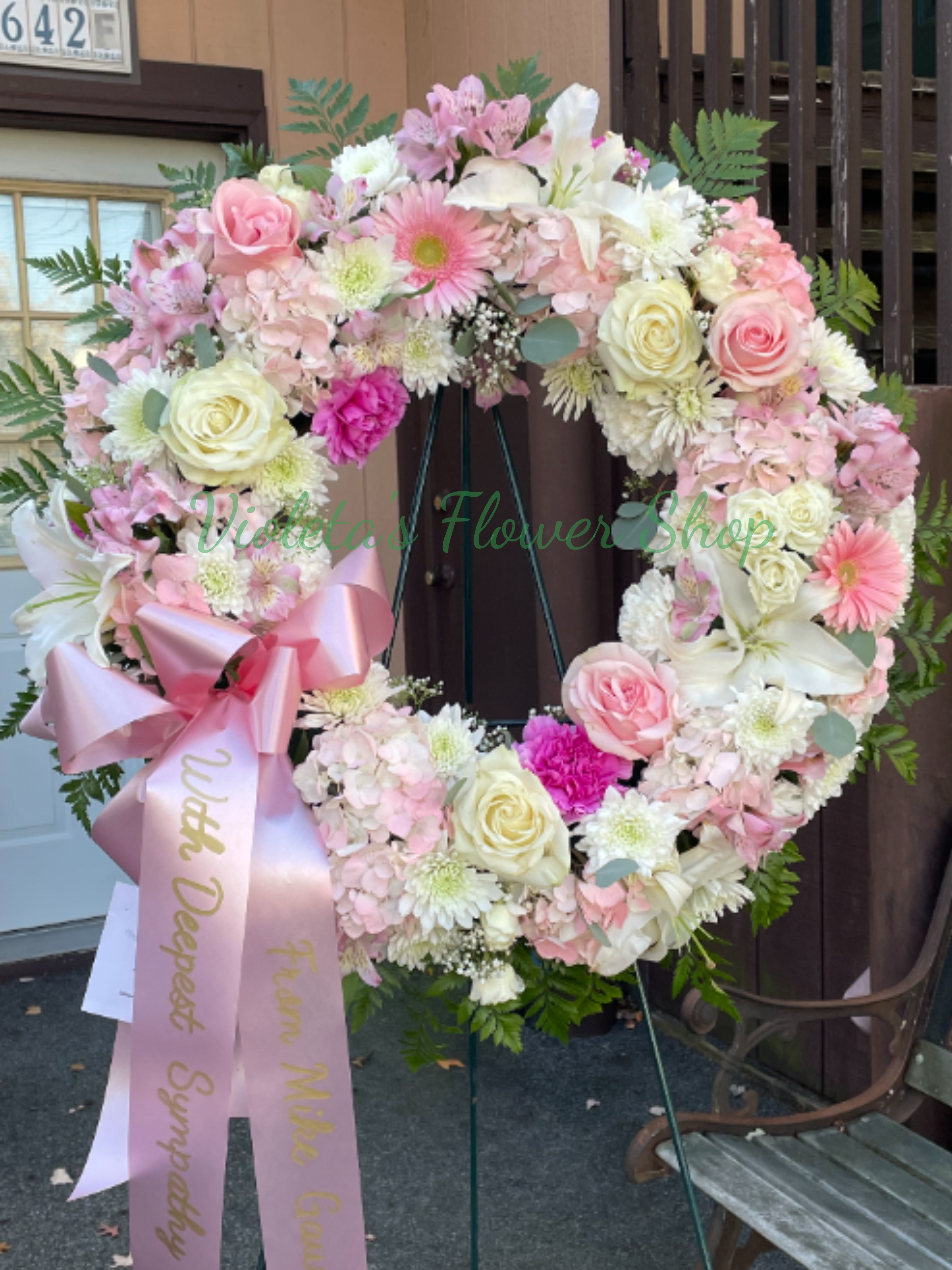 Always Remembered Wreath - A soft pink and white sympathy wreath arranged on a sturdy metal easel offers a heartfelt way to honor a life. Ideal for a funeral or memorial service, this large circular or heart-shaped flower arrangement reads as a formal tribute-romantic and sentimental-so the person you're remembering will be surrounded by gentle color and thoughtful detail.  The palette of pink, white, green, and touches of purple creates a calming, graceful look. A wide pink satin ribbon with gold lettering is draped from the lower left for a traditional memorial presentation; a small card can be included to carry your personal message.  