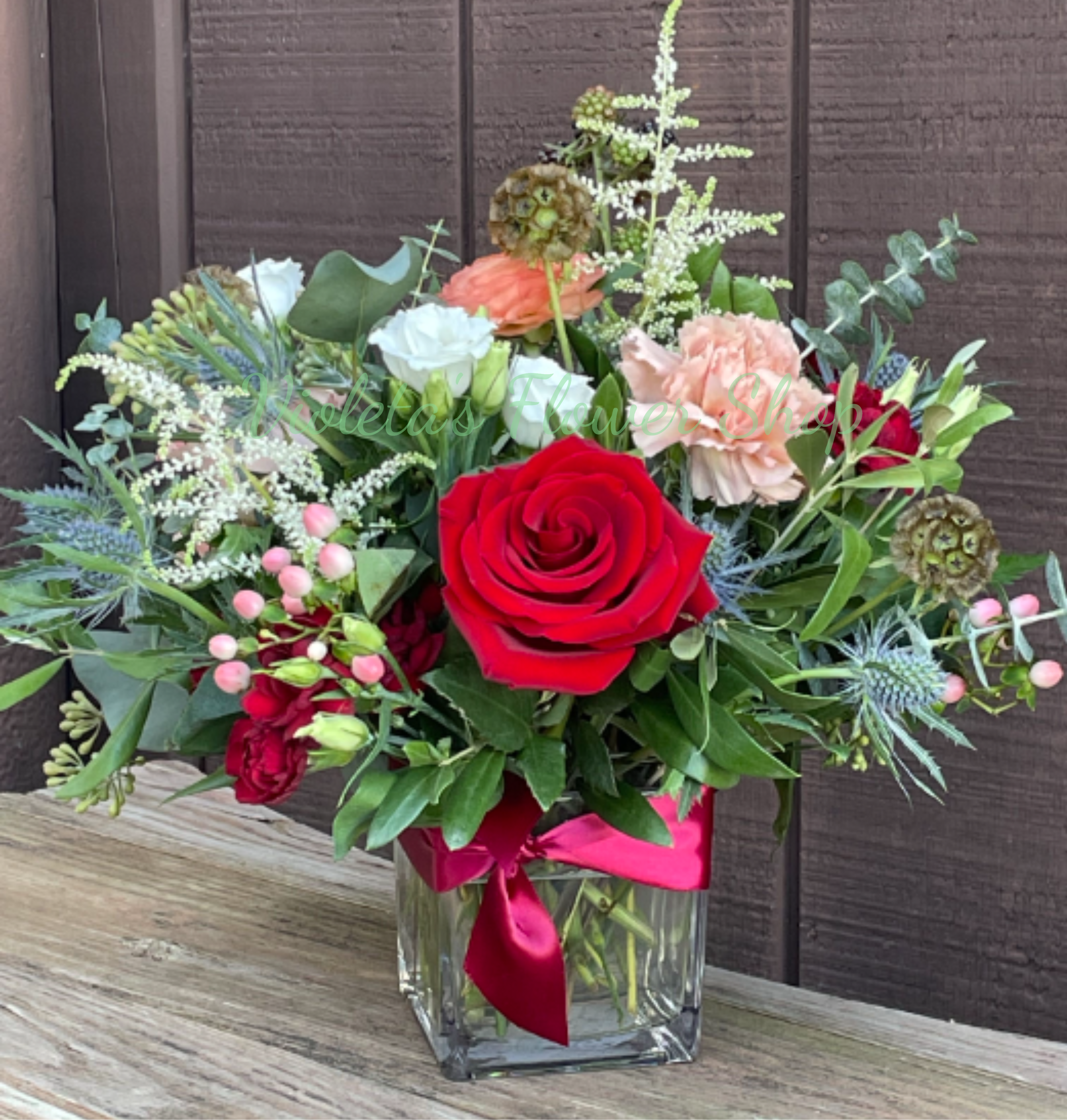 Fierce Romance Arrangement - A bold red rose anchors this romantic anniversary arrangement, surrounded by blush pink and crisp white blooms for a garden-fresh look that reads "love and romance" at a glance. Perfect for anniversaries, a retirement celebration, or a "just because" surprise, the composition balances classic roses and spray roses with soft lisianthus and carnations so the recipient feels truly seen and celebrated.  Textural accents-berry clusters, spiky blue thistle-like blooms, and airy white filler-create gentle movement through the rounded, hand-arranged bouquet, while varied greenery adds depth and contrast. The flowers arrive arranged in a clear square glass vase and finished with a bright pink satin ribbon for a polished, gift-ready presentation that still feels personal and hand-tied.  
