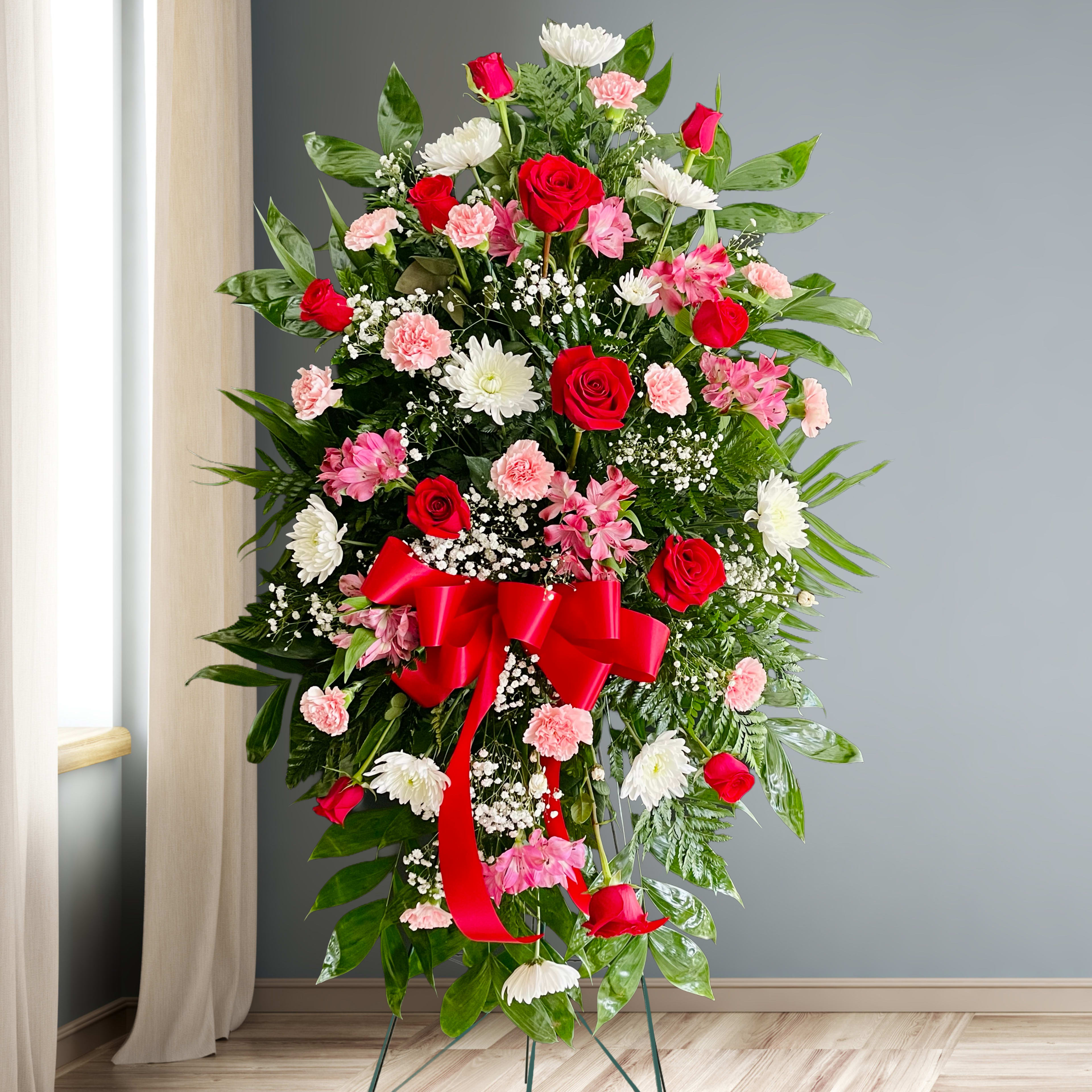 Standing floral spray with red roses, pink carnations, and white daisies on a red ribboned easel