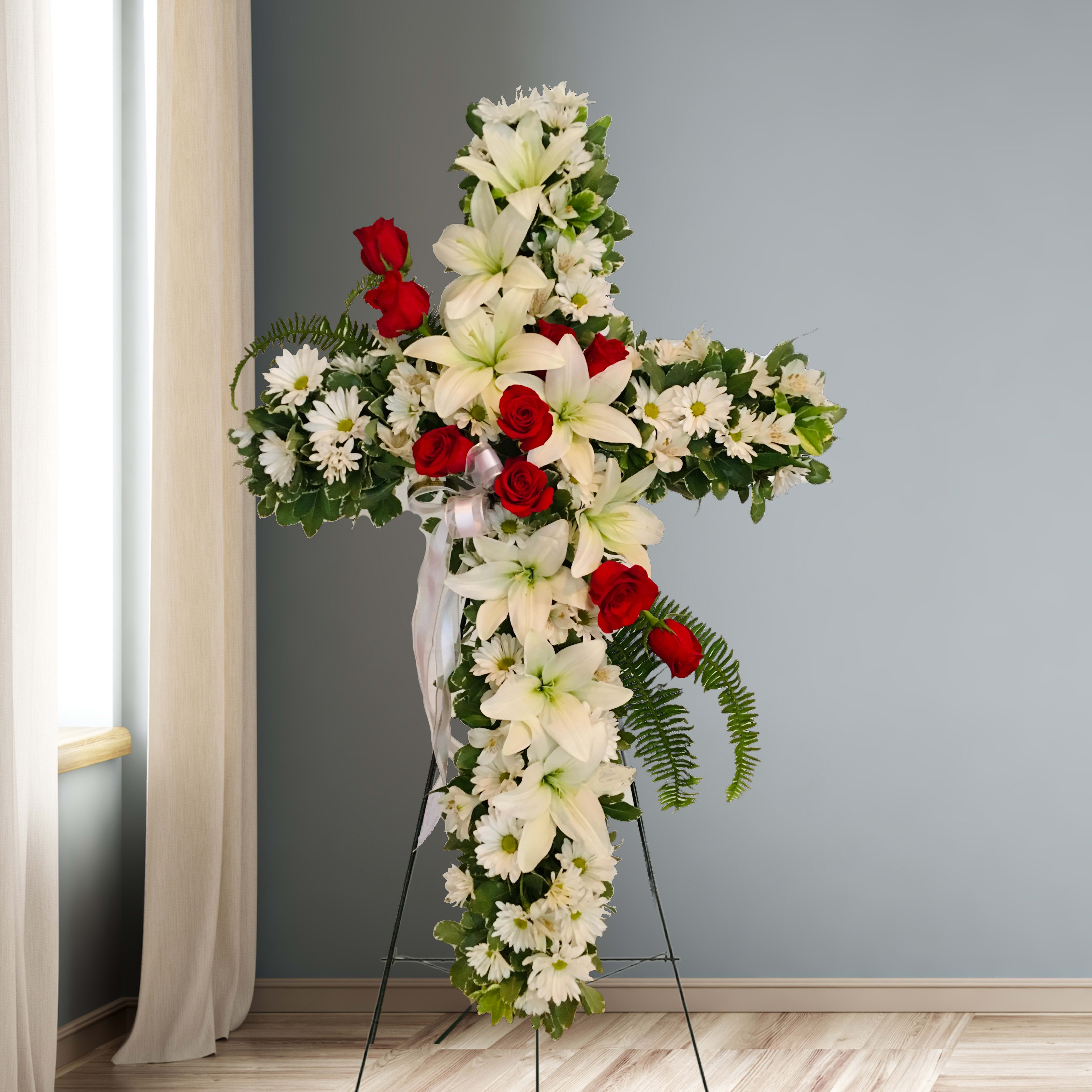 Cross-shaped floral tribute with white lilies, daisies, and red roses on a stand