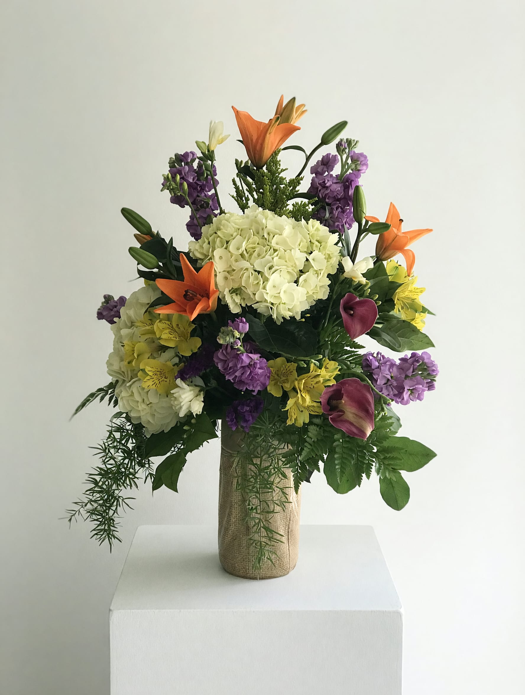 Burlap Memories - A lush flower arrangement that says, She's a Cool Mom. Surrounded by bright orange lilies, cheerful yellow alstroemeria, purple stock-like spikes and deep burgundy calla lilies for contrast. As a general occasion bouquet-perfect for Mother's Day, a birthday, or "just because" celebrations-this upright, full design reads lively and celebratory, the color mix sending a message of warmth, admiration, and playful confidence to the person you're honoring.  The composition layers blooms at different heights for a generous, abundant silhouette that feels full from every angle. Textures range from the soft, cloudlike hydrangea to the sleek calla lilies and spiky purple stems, all presented in a rustic burlap-wrapped vase that complements the vivid palette. Dimensions and exact flower counts vary by size selection; each arrangement is hand-crafted to maintain the same lush look and color balance.  Give this to a mom, friend, or mentor who loves bold color and relaxed elegance-the kind of bouquet that will make her feel celebrated and seen. Care is simple: keep the vase filled with fresh water and refresh every few days; for same-day or scheduled delivery, select your preferred date at checkout.