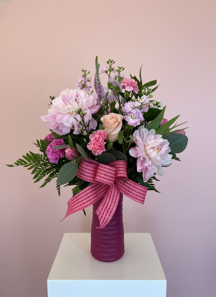 Sweet Pink - Soft blush peonies anchor this Sweet Pink flower arrangement, making it a thoughtful birthday bouquet or heartfelt Welcome Baby Girl gift that reads as joyful and intimate. The rounded, full composition blends a peach rose, pink carnations, lavender stock and other delicate filler blooms so the person you're thinking of will feel genuinely celebrated the moment they see it.  Pinks and purples are softened by white accents and deep green foliage for a balanced, feminine palette. The stems are arranged in a magenta ceramic vase finished with a wide striped ribbon for an instantly gift-ready presentation; textures range from the peonies' lush petals to the airy stock and crisp greenery, giving the arrangement visual depth and scent-rich appeal.  Ideal for New Baby Girl, Birthdays, or "Just Because" moments, this arrangement suits someone who loves soft, elegant color stories and a classic, hand-tied look in a keepsake vase. Order with confidence for delivery to make her feel truly seen; simple care-fresh water and a cool spot away from direct sun-will help the blooms last.
