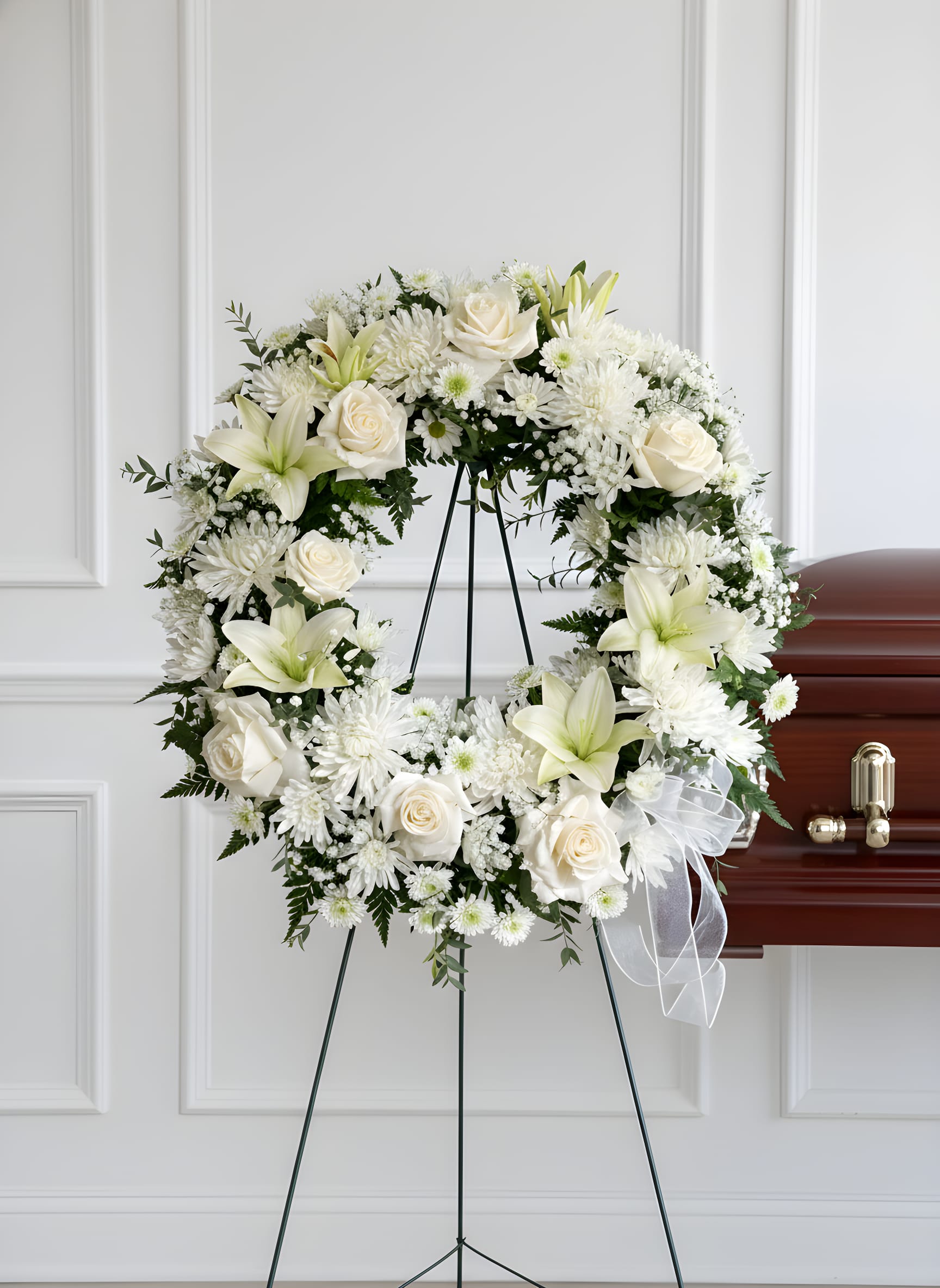 Serene Remembrance - A large white funeral wreath arranged on a standing easel, designed to offer a serene and traditional tribute for a sympathy or funeral service. The circular wreath of white roses, lilies, chrysanthemums and small daisy accents creates a calming, respectful presence beside the casket, while the white ribbon adds a soft memorial touch-perfect for expressing sympathy, honoring a life, or sending condolences to the family.  Dense dark green foliage frames the blooms for contrast, emphasizing the bright whites and the wreath's formal silhouette. The mix of textures-from velvety rose petals to the trumpet shape of lilies and the cushiony chrysanthemum blooms-gives the arrangement visual depth and a gentle, comforting feel. Included with a floral wreath stand and finished with a white ribbon, this arrangement is sized and styled to stand gracefully beside a polished casket at a funeral service, communicating sympathy and reverence without words.