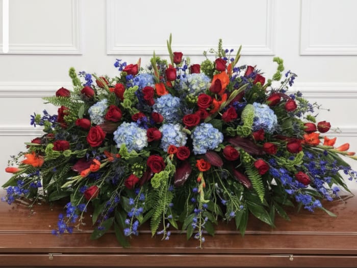 Last Sunset - A large, low casket spray designed for a sympathy or funeral tribute that commands attention with its vivid red, blue, and orange palette. The mix of red roses and deep blue hydrangeas creates a strong, ceremonial contrast while the orange lilies add warmth and a sense of depth-the kind of arrangement that helps the person you're honoring feel remembered and celebrated in a dignified way.  This full, formal spray sits low and spreads broadly across the casket surface, with dense clusters of roses and hydrangeas balanced by cascading blue accents and abundant greenery. The orange lily blooms punctuate the composition, offering bright highlights against the rich reds and blues; the overall texture is lush and layered, conveying care and permanence appropriate for a memorial setting.  Choose this spray when you want a bold, respectful expression of sympathy that reads clearly from a distance and feels intimate up close. Sized to cover a large casket area, it arrives ready to place and benefits from fresh water at delivery; for specific sizing or personalization requests, please contact us to ensure the tribute matches your intentions.
