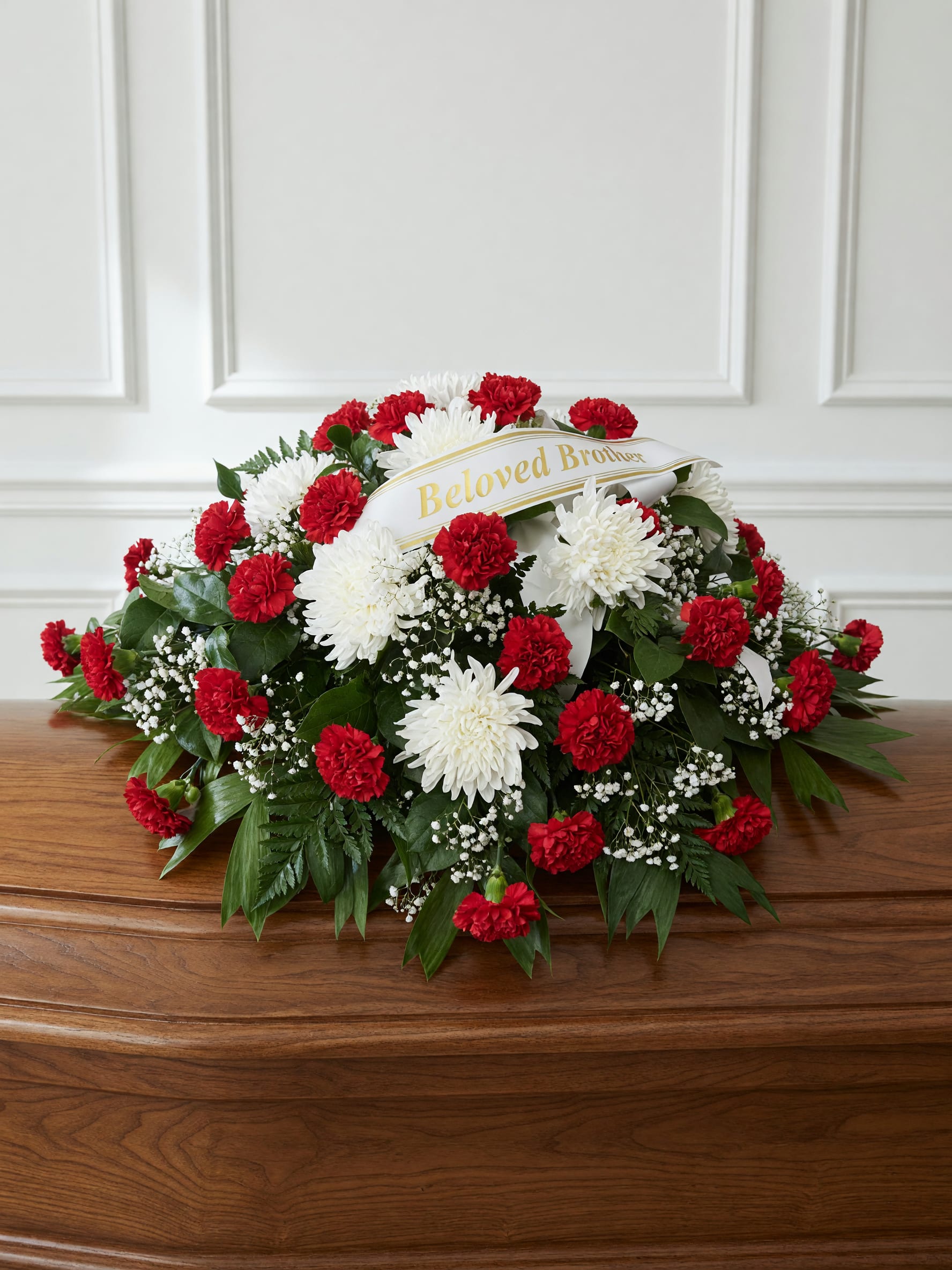 Beloved  - A low, oval sympathy spray of deep red carnations and pure white chrysanthemums makes a respectful, classic memorial statement for a funeral or remembrance service. This funeral arrangement, intended as a tribute for a brother, pairs the formal red-and-white palette with soft accents of baby's breath and mixed greenery to create a comforting, dignified presentation that reads clearly from any pew or gathering space.  The flowers are densely clustered into a full, rounded oval that lies low for placement on a casket or memorial table; textures range from the ruffled carnation petals to the crisp chrysanthemum blooms and delicate baby's breath. A white funeral customizable ribbon is included, reinforcing the message of love and remembrance. The red, white, and green color scheme offers strong contrast and a timeless look appropriate for traditional services.  This spray is meant to convey heartfelt sympathy and honor - the kind of tribute that helps family and friends feel seen in their grief. It arrives ready for display; please let us know any specific wording or delivery details when ordering to ensure it reaches the service on time.