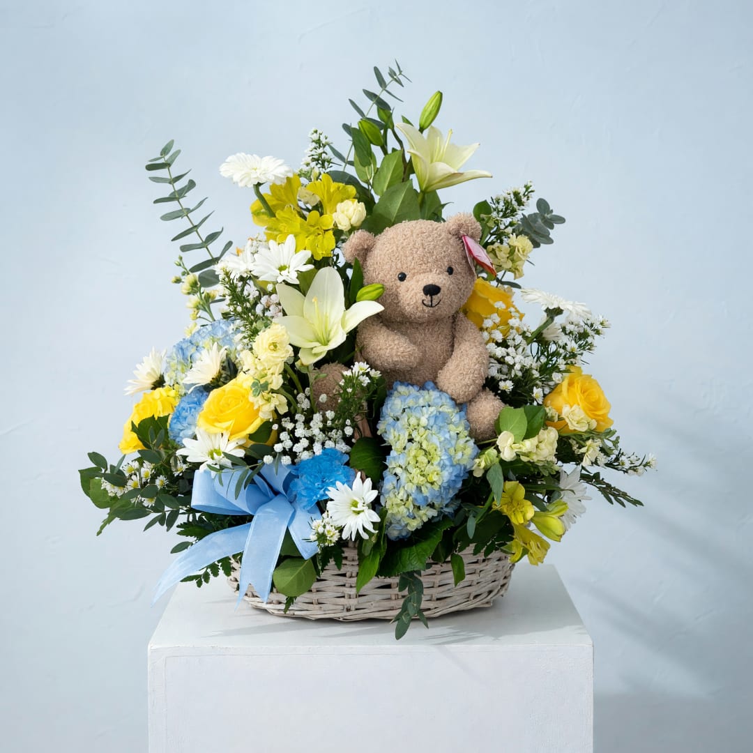 Basket arrangement with a teddy bear and mixed flowers in blue, yellow, and white