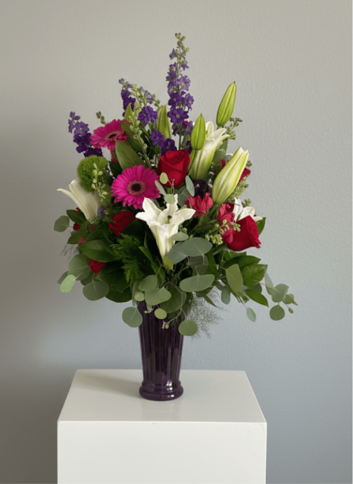 Daz Luz - A vivid, celebratory mixed bouquet that makes the recipient feel truly noticed, "Daz Luz" or "you give light" is a flower arrangement built to impress for birthdays, anniversaries, or romantic gifting. The tall purple glass vase and upright silhouette give this arrangement a slightly formal, sophisticated presence that still reads joyful; it's the kind of gift that announces the moment and makes someone feel celebrated.  This full design layers red roses, white lilies, bright pink gerbera daisies, purple spike flowers, snapdragons and alstroemeria amid rounded eucalyptus foliage for a lush, balanced look. The rich red, crisp white, playful pink and deep purple palette creates strong contrast and depth, while the tall vase accentuates the arrangement's vertical lines. The glass vase provides a clean, modern finish and keeps stems visible for a fresh presentation.  Perfect for the person who deserves something bold-a romantic partner on an anniversary, a friend's milestone birthday, or a hostess you want to honor, Daz Luz reads as both festive and refined. Arrives ready to display in the included purple glass vase; change water every 2-3 days, recut stems at an angle, and keep away from direct heat to prolong bloom life.
