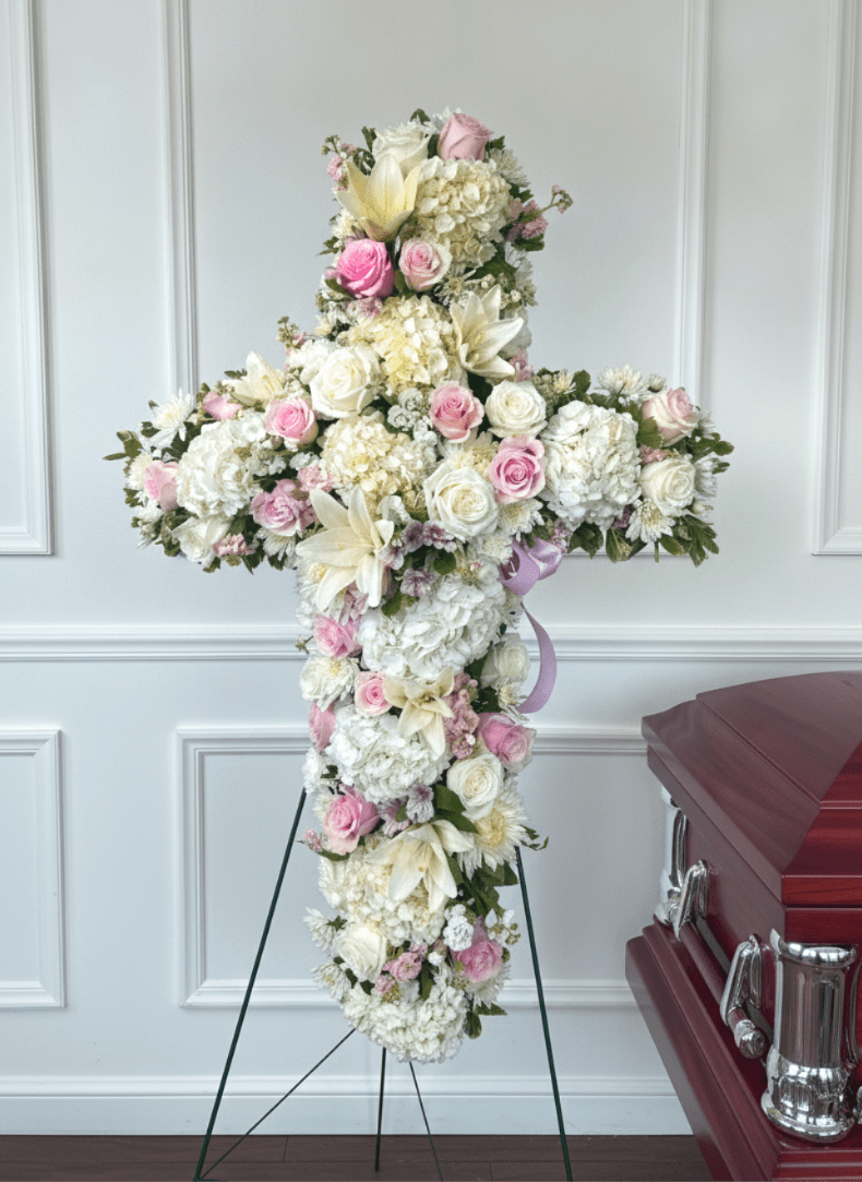 Cross of Hope - A white and blush cross-shaped funeral arrangement arranged for a service or memorial, designed to offer a quiet, reverent presence that honors the life being remembered. This sympathy cross is meant for a funeral or graveside service and presents a calming palette-soft whites, pale pinks, and touches of lavender and green-to convey comfort and respect to the person you're remembering.  Densely covered with large white hydrangea clusters, pale roses, white lilies, daisies and mixed pastel blooms, the design reads formal and polished. The cross is finished with a lavender ribbon and mounted on a metal easel stand or floral stand for upright display; it can also be placed near a casket as part of a service. The tightly packed flowers create a full, tactile surface that reads from a distance and close up-the hydrangeas give volume, the roses add gentle romance, and the lilies bring classic, clean lines.  Made to sit prominently beside a casket or at a memorial space, this arrangement is a meaningful way to say you're keeping someone in your thoughts and prayers. Size and exact stem counts are crafted to suit service needs; please let us know the service location and desired stand option when ordering so we can ensure proper delivery and presentation.