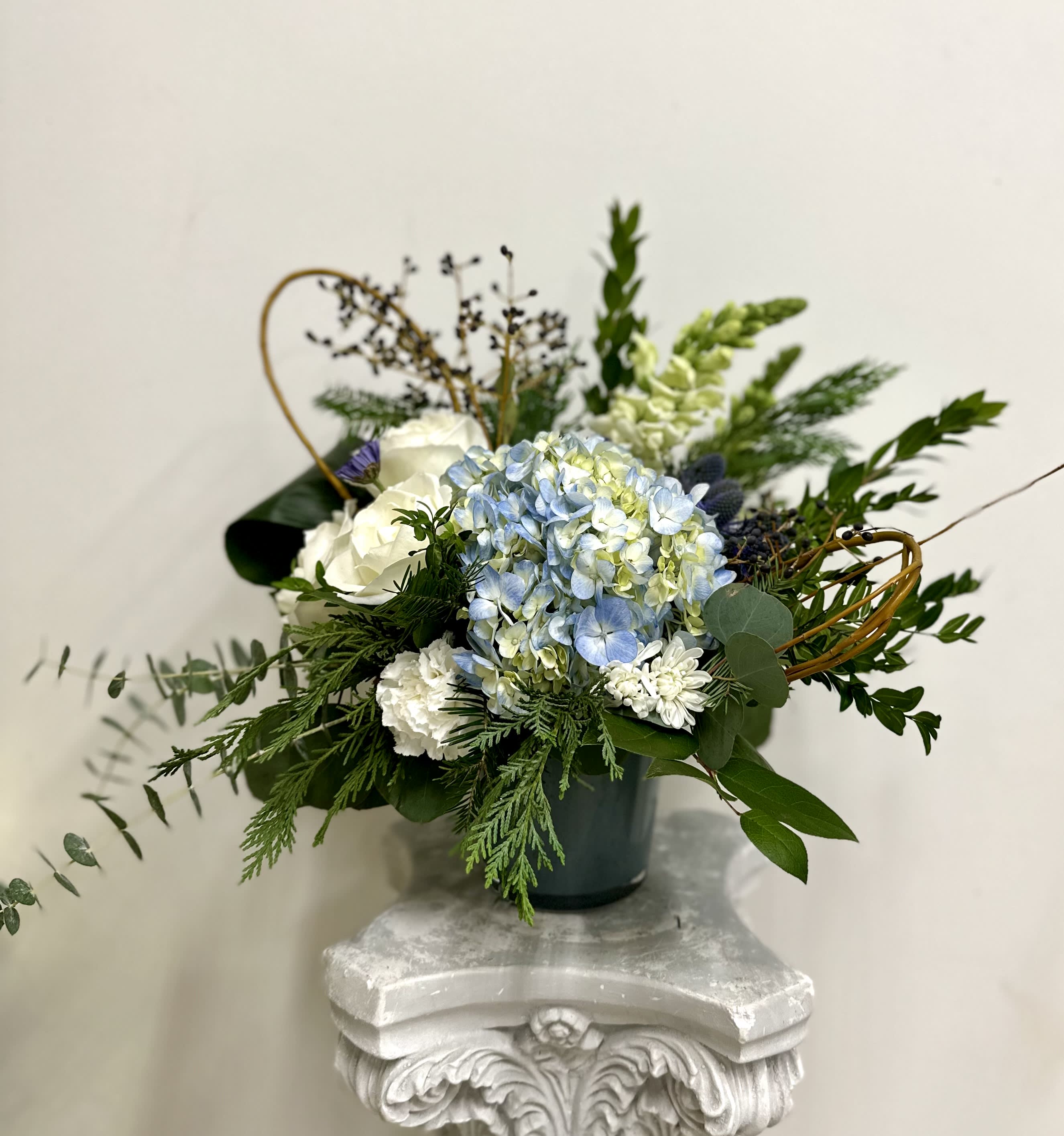 Winter Blues by Westford Florist Art by Veny - Embrace the serene beauty of winter with Winter Blues by Westford Florist Art by Veny. This exquisite arrangement features a harmonious blend of blue hydrangeas, elegant white roses, and whimsical snapdragons, all artfully showcased in a stunning dusty blue glass container. The rich combination of thistle and delicate berries adds depth to this modern floral design, while plush cushions and charming carnations bring warmth to any space. Perfect for brightening up your home or as a thoughtful gift, Winter Blues captures the essence of the season's tranquility and elegance. Elevate your environment with this sophisticated centerpiece that speaks to both style and serenity.