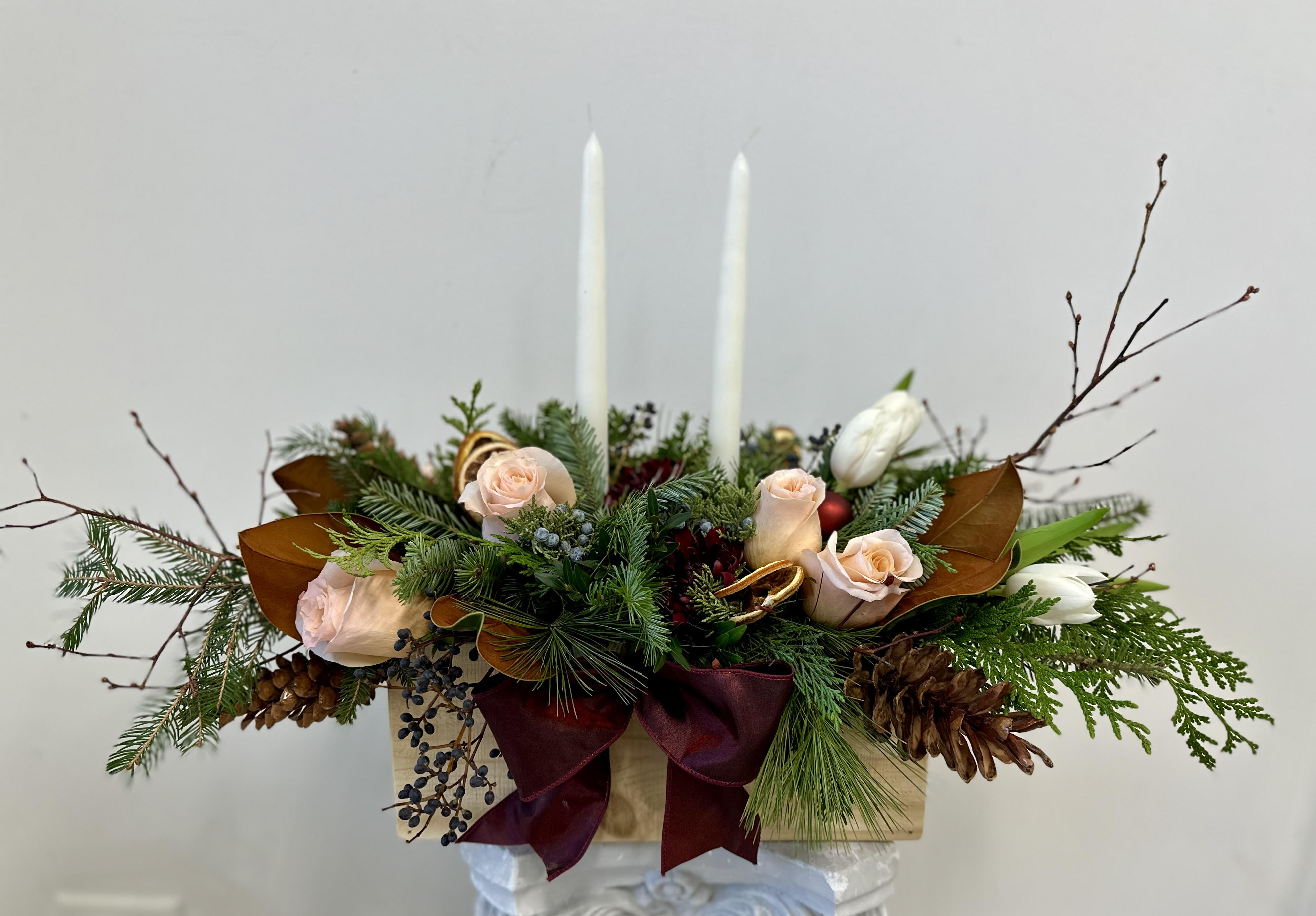 Candlelight Grande Centerpiece by Westford Florist Art by Veny - Transform your festive gatherings into unforgettable moments with the Candlelight Grande Centerpiece by Westford Florist Art by Veny. This exquisite arrangement combines a thoughtful mix of seasonal flowers and natural elements, infusing your space with rustic elegance and cheer. Set in an all-natural, untreated wooden box, this centerpiece captures the essence of holiday warmth through its vibrant palette of festive tones. The freshness of greenery not only enhances its visual appeal but also invites the spirit of the season indoors. Ideal for any table setting, let the Candlelight Grande be the heart of your celebrations, sparking joy and conversation at every occasion. Two (2) candle lights are included.