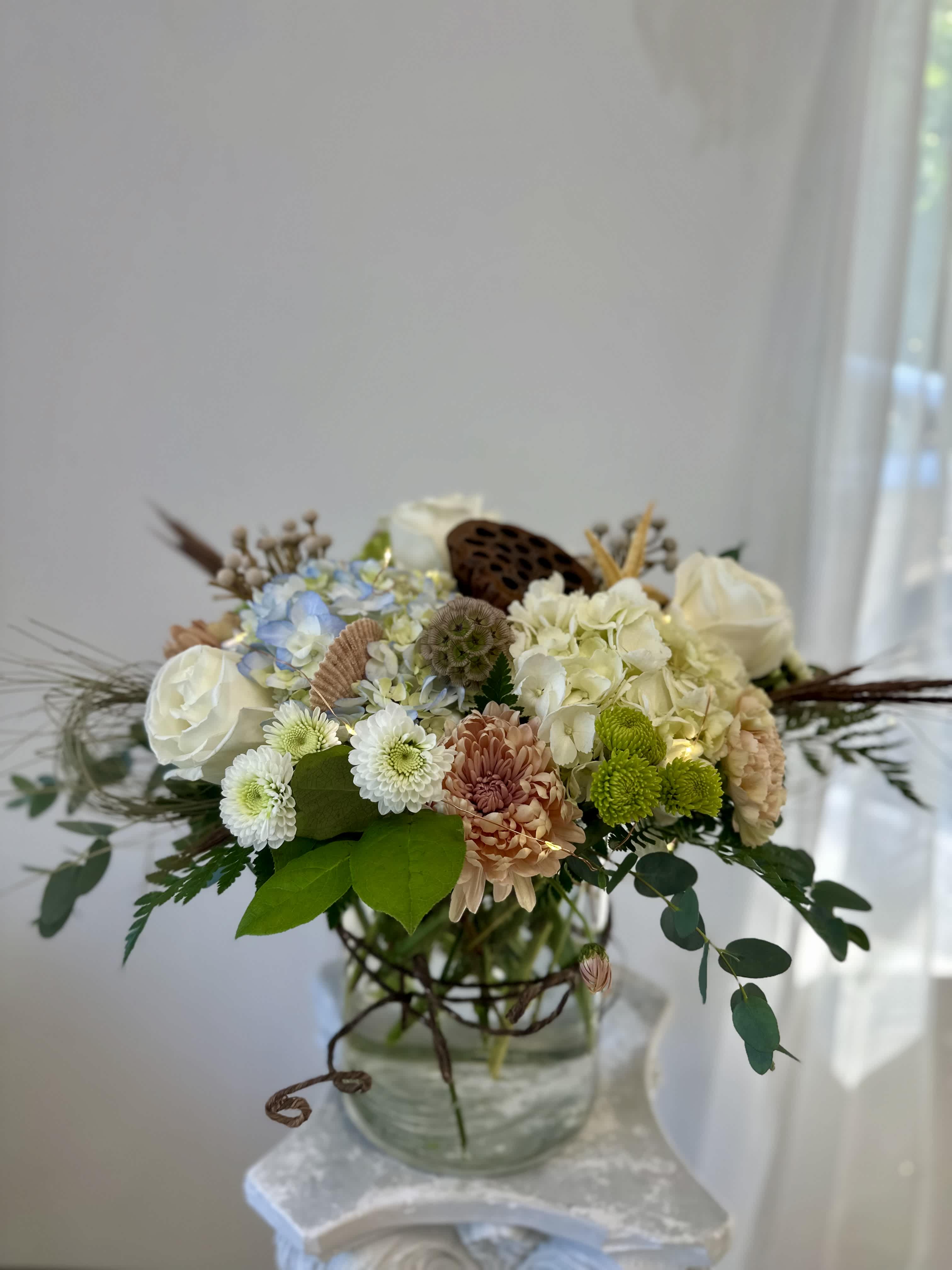Ocean Breeze by Westford Florist Art by Veny - Transform your space into a serene oasis with Ocean Breeze by Westford Florist Art by Veny. This stunning floral masterpiece encapsulates the calming essence of the sea, expertly designed to infuse your home with tranquility. Each arrangement features an exquisite blend of hydrangeas, roses, poms, and brunia flowers, complemented by vibrant green trick and delicate peach carnations. Accented with natural shells and pods alongside tasteful dry weeds, this enchanting collection is further enhanced by subtle LED lights that bring a warm glow to any setting. Elevate your decor and create an inviting atmosphere that soothes the senses—Ocean Breeze is more than just an arrangement; it’s a coastal retreat for your home. Experience the calming effects of this exceptional arrangement by ordering now from Westford Florist. Our talented team will carefully handcraft each piece using only the freshest and highest quality flowers available. Every stem is selected with great care so that each time you gaze upon Ocean Breeze; it transports you to sandy beaches and clear blue waters. Immerse yourself in the tranquil beauty of the ocean with Ocean Breeze by Westford Florist. Let its stunning mix of florals and natural elements transport you into a world where serenity reigns supreme. Order today and elevate your surroundings with the undeniably beautiful Ocean Breeze.