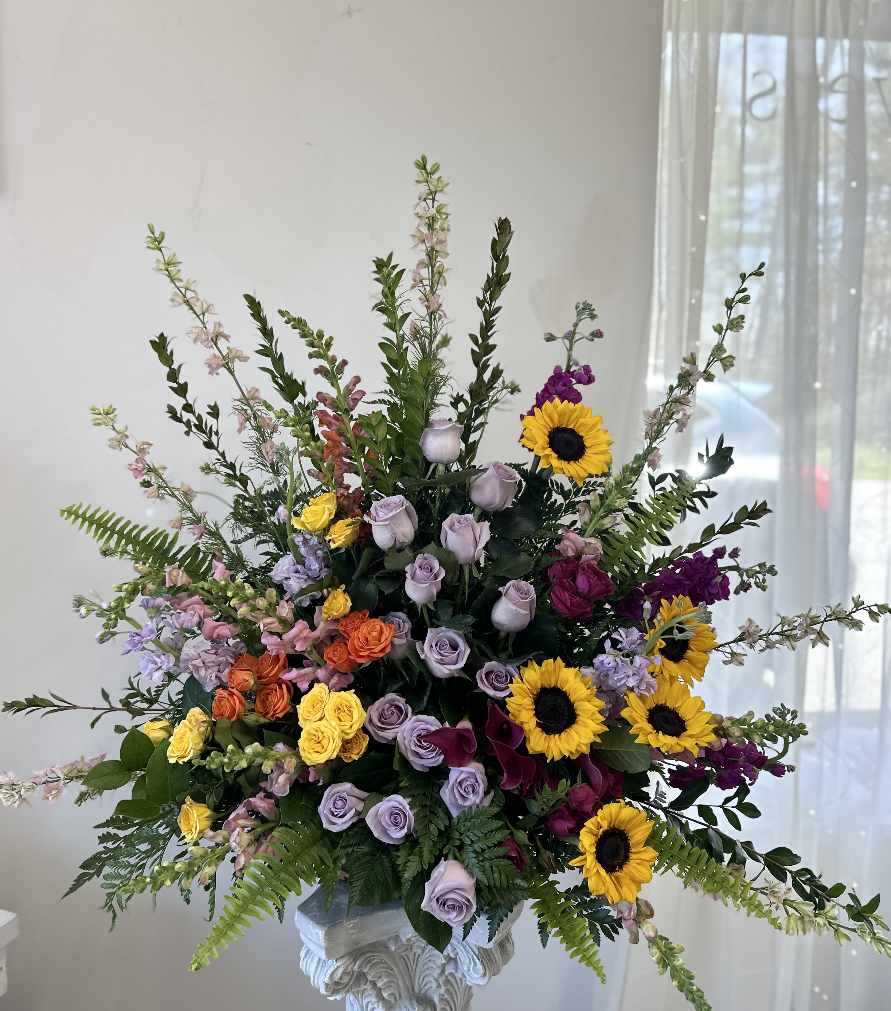 Large mixed floral arrangement with sunflowers, roses, and purple blooms in a pedestal vase