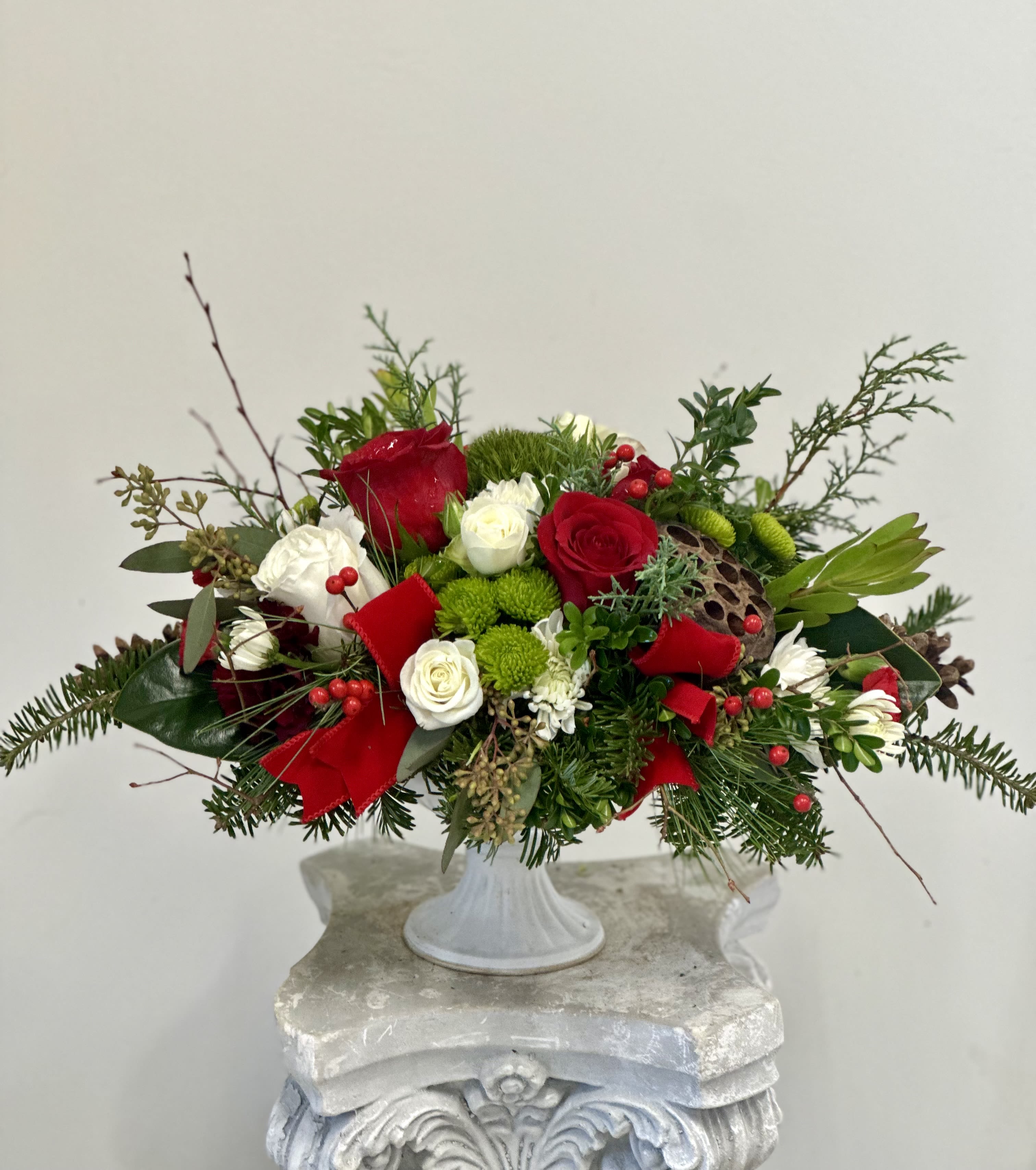 Holiday Harmony Centerpiece by Westford Florist Art by Veny - Elevate your holiday gatherings with the Holiday Harmony Centerpiece by Westford Florist Art by Veny. This exquisite arrangement features a stunning blend of white and red roses and white spray roses, complemented by cheerful green and white poms, green trick and red and burgundy mini carnations. Nestled among vibrant safari sunset blooms and festive red berries, lotus pod, natural pinecones and magnolia leaves each element is thoughtfully curated to bring warmth and joy to your celebrations. Housed in a stylish tall pedestal metal container topped by bright velvet ribbon, this centerpiece exudes sophistication while celebrating the beauty of the season. Natural pinecones and lush Christmas greenery complete the design, creating a captivating focal point that enchants both family and guests alike. Make this holiday season truly memorable with an elegant touch that embodies harmony and festive spirit.