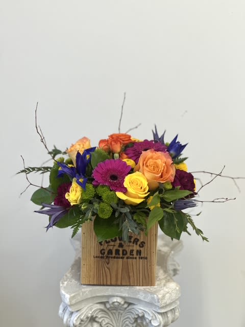 Bright mixed arrangement of yellow and orange roses, purple gerbera and blue iris in a wooden box.