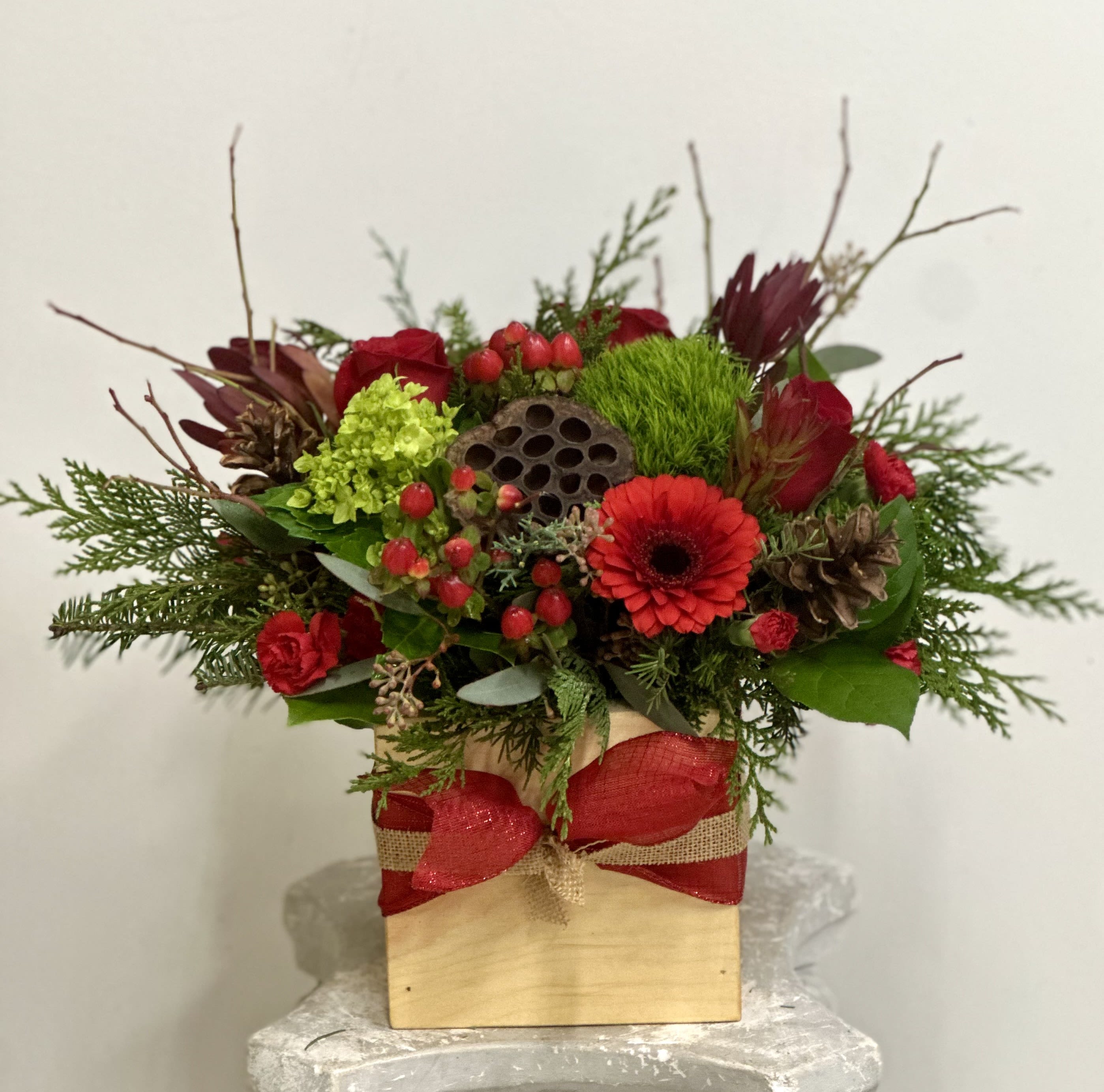 Heritage Kindle by Westford Florist Art by Veny - Elevate your space with the Heritage Kindle by Westford Florist Art by Veny, a stunning arrangement that beautifully captures the essence of nature's bounty. This exquisite centerpiece features a delightful blend of vibrant red and lush green flowers, harmoniously accented with pinecones, safari sunset blooms, berries, and rich evergreens. Each element is thoughtfully nestled in a handcrafted rustic wooden box made from aromatic red cedar, exuding warmth and charm. The elegant ribbon adornment enhances its sophisticated appeal while ensuring it fits seamlessly into any décor. Ideal for gifting or as a striking focal point in your home or office, the Heritage Kindle brings together artistry and natural beauty in one captivating piece. Experience the perfect blend of tradition and elegance—transforming ordinary moments into cherished memories.