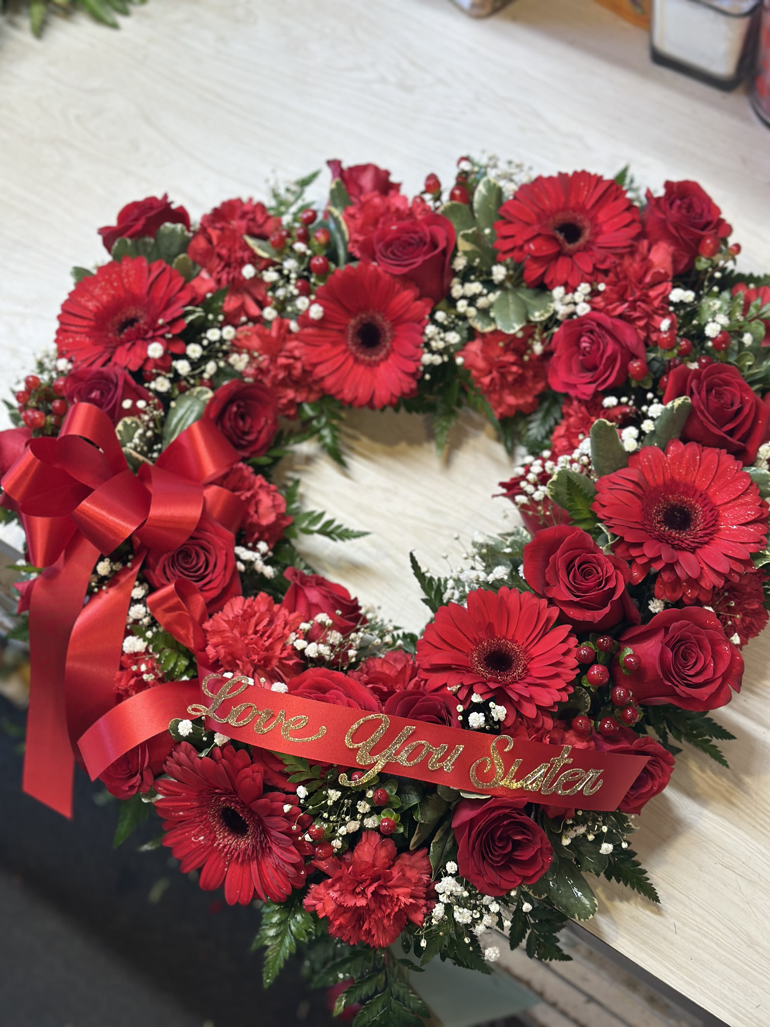 Crimson Heart Tribute - A striking open-heart arrangement designed to honor deep love and remembrane. Featuring rich red roses, vibrant red gerbera daisies, and lush carnations, this piece is beautifully accented with delicate baby's breath.  