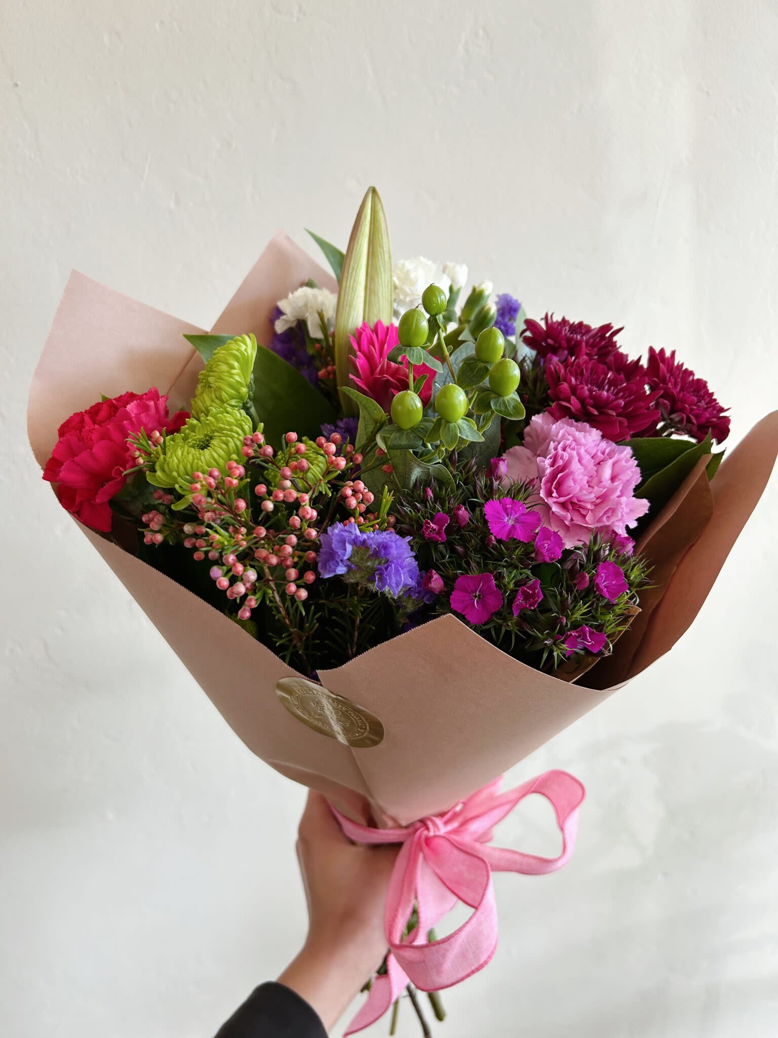 Wrapped Seasonal Bouquet - Bright, garden-style color takes center stage in this wrapped seasonal bouquet - a lively mixed-color flower arrangement perfect for a birthday, anniversary, or a Mother's Day surprise. The tall lily bud rising from the center gives the bouquet a dramatic vertical focus while pink, magenta, purple and lime-green blooms combine to create an uplifting, slightly romantic impression the person you're thinking of will notice the moment they receive it.  This hand-tied bouquet features lilies, carnations, chrysanthemums and dianthus with small berry-like accents for added texture, arranged loose and abundant for a natural, celebratory feel. It's wrapped in soft blush or beige paper and finished with a pink satin ribbon, giving a polished yet fresh presentation that reads as both thoughtful and joyful. The mix of ruffled and clustered blooms creates a full, layered shape that photographs beautifully and smells subtly floral thanks to the lilies.  Ideal for marking anniversaries, birthdays, or simply saying "I'm thinking of you" on Mother's Day, this bouquet comes ready to gift and is sized for easy carrying and display in a simple vase. To keep the blooms looking their best, trim stems and refresh the water daily; with basic care the arrangement will continue to brighten a room for days.
