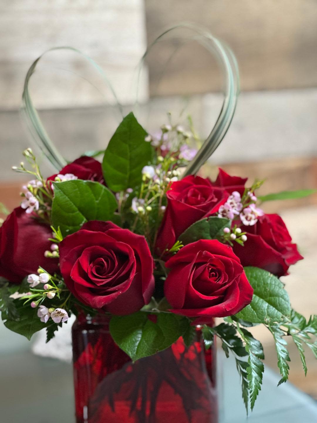 Short arrangement of red roses with small pink blossoms in a red glass cube vase with a heart-shaped grass accent.