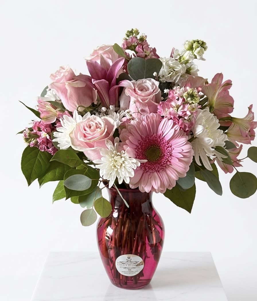 The Blushing Luxe - Elevate your space with "The Blushing Luxe," a professionally designed, mixed-flower arrangement that marries bold color with elegant textures. This striking centerpiece features perfectly formed pink Gerbera daisies—the stars of the show—with no missing petals, creating a full and uniform look. They are artfully layered with rich, deep-hot pink  carnations, delicate light-pink carnations, and pristine white chrysanthemums, all set among lush green foliage.