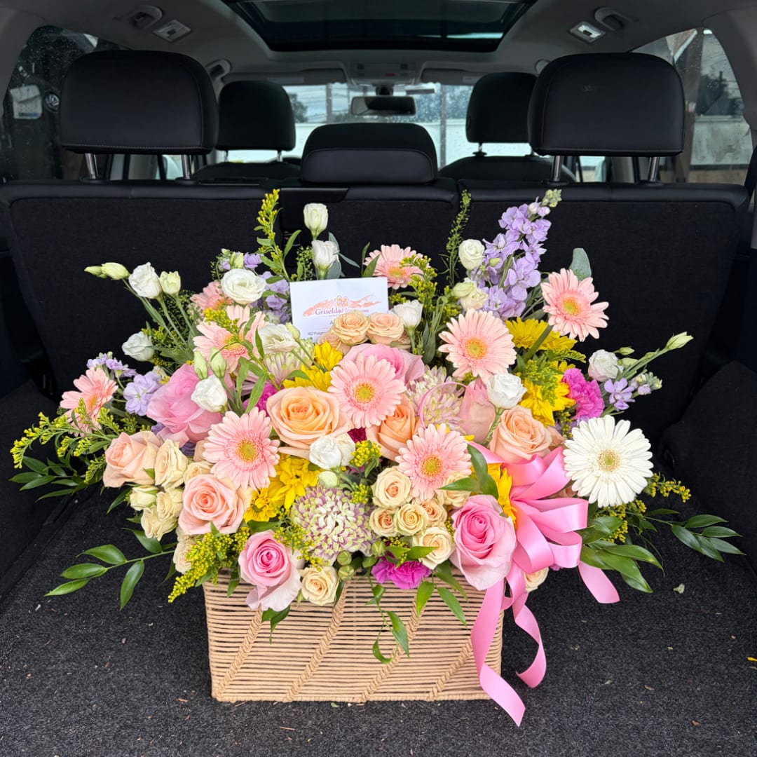 Large mixed flower arrangement in a woven basket with a pink ribbon