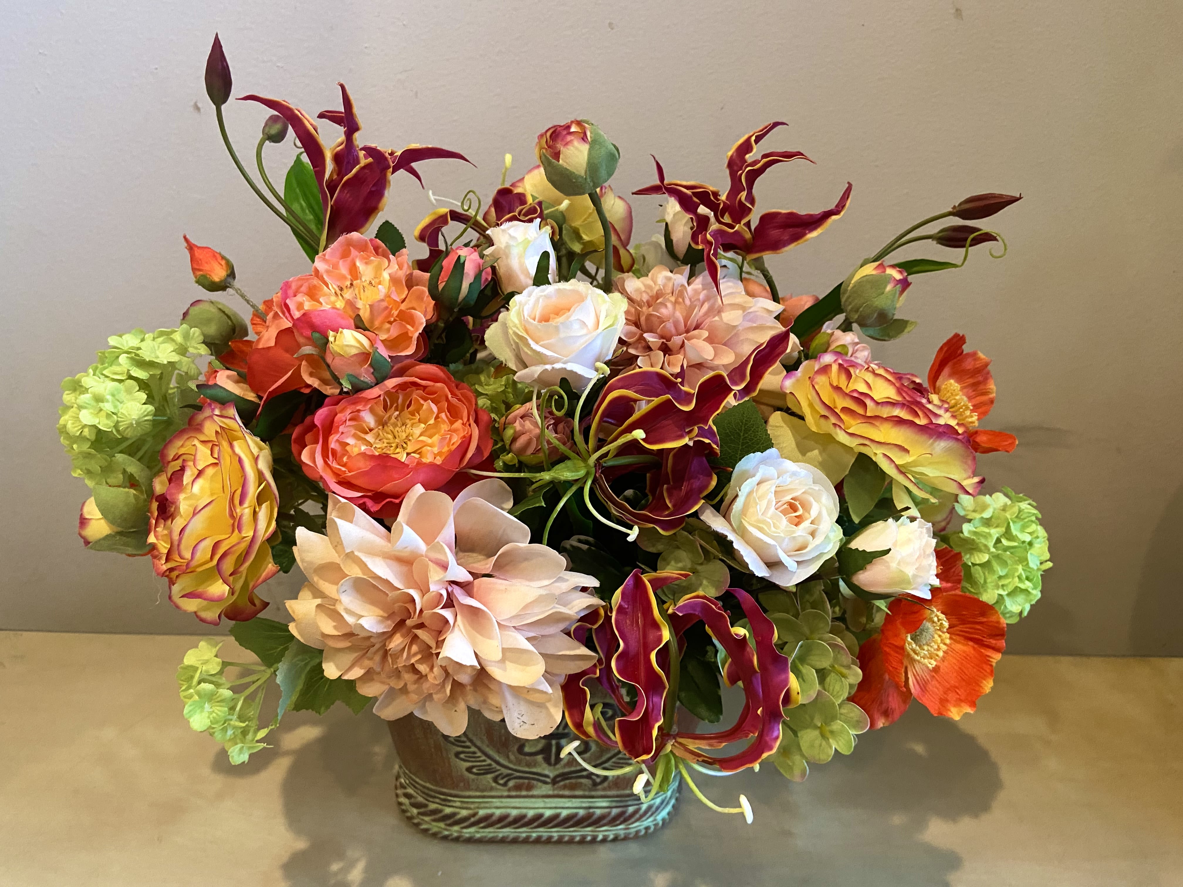 Lush centerpiece of orange, peach, and yellow blooms with burgundy accents in a decorative ceramic container